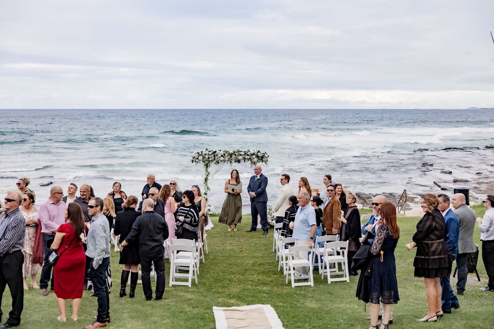 Outdoor coastal wedding ceremony setup with white chairs and guests gathering on a grassy cliff overlooking the ocean.