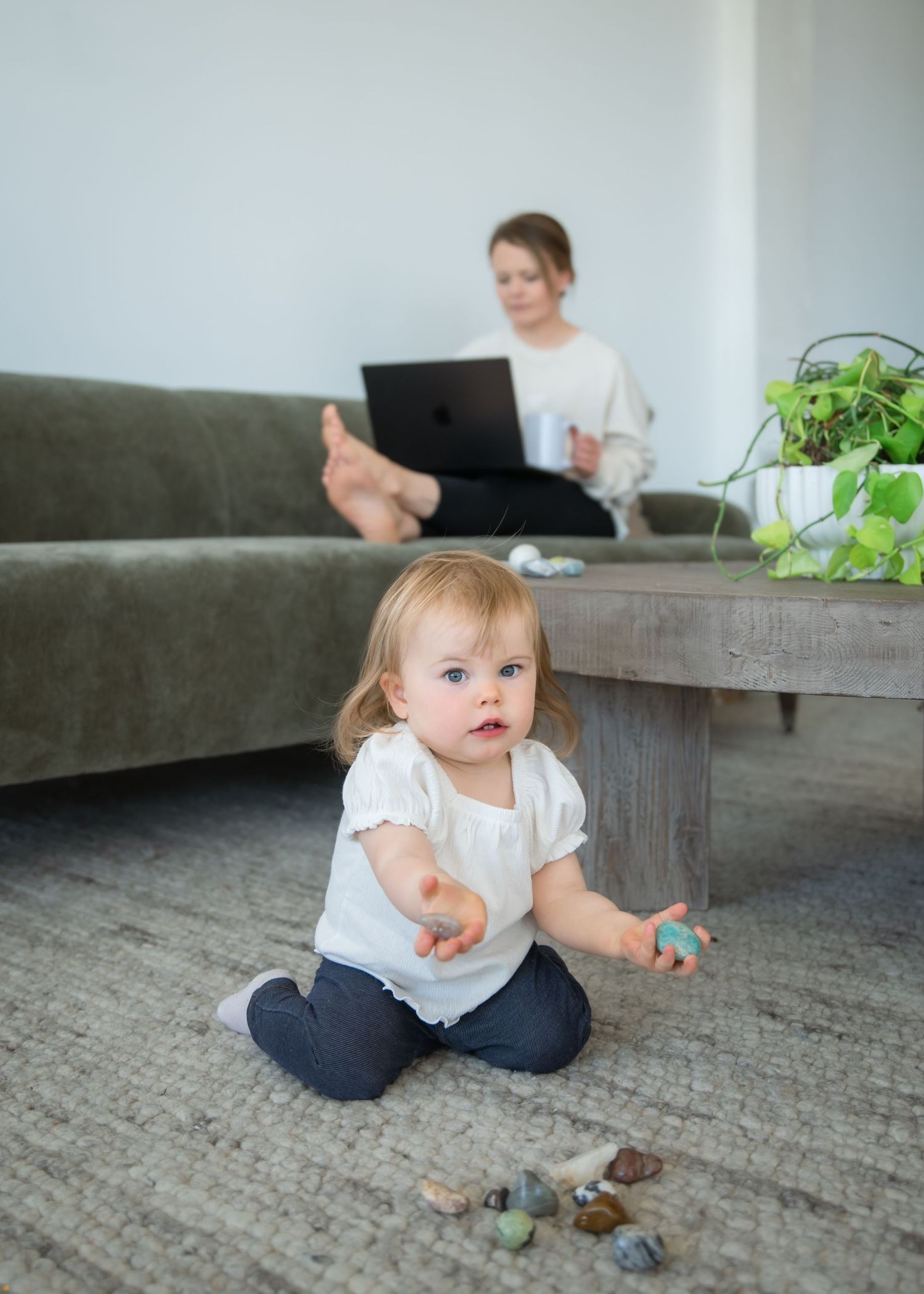 Small child playing on floor with toys while parent works from home on laptop sitting on grey couch.