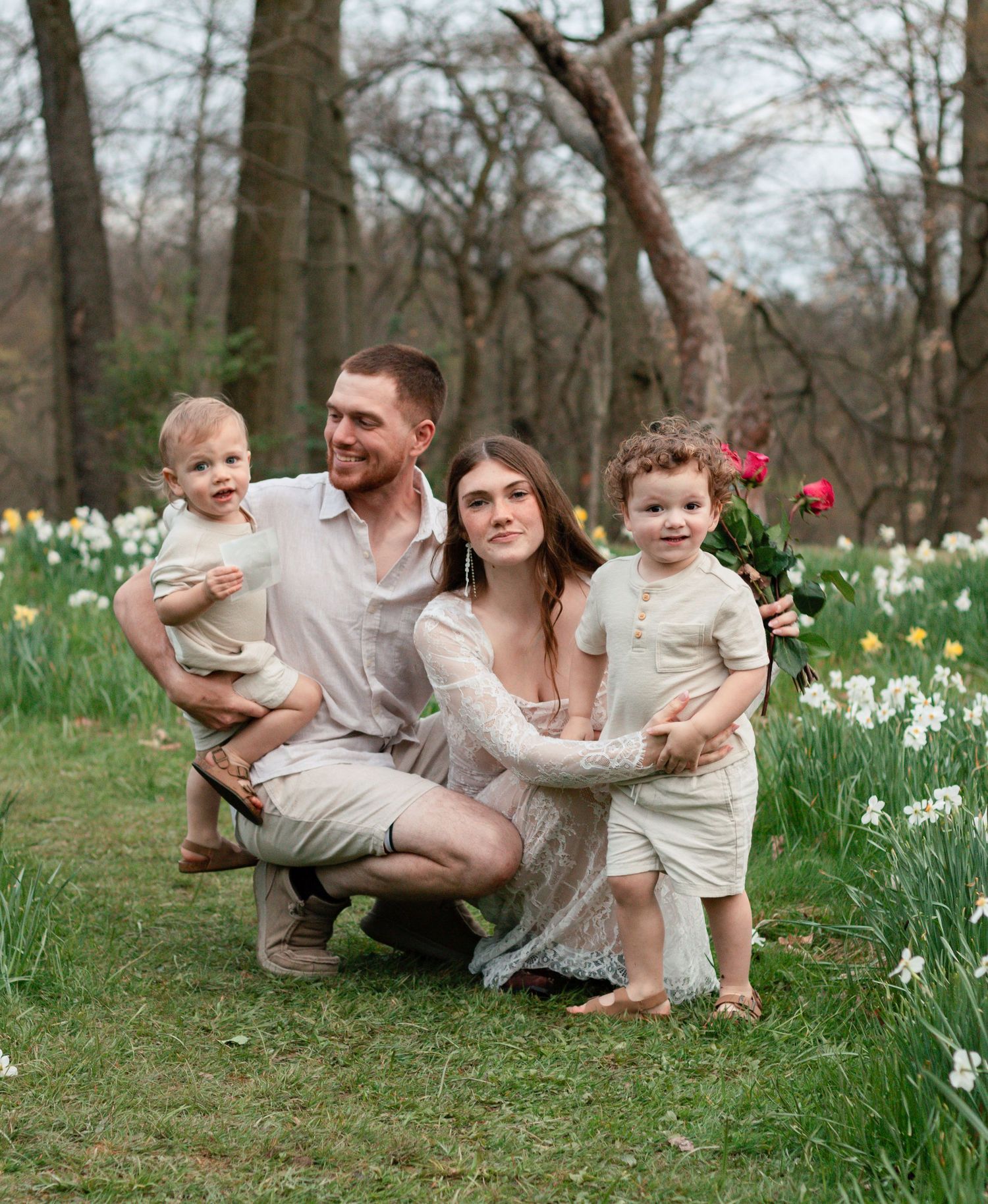 Family wearing white and beige outfits poses outdoors in a spring garden with blooming daffodils.