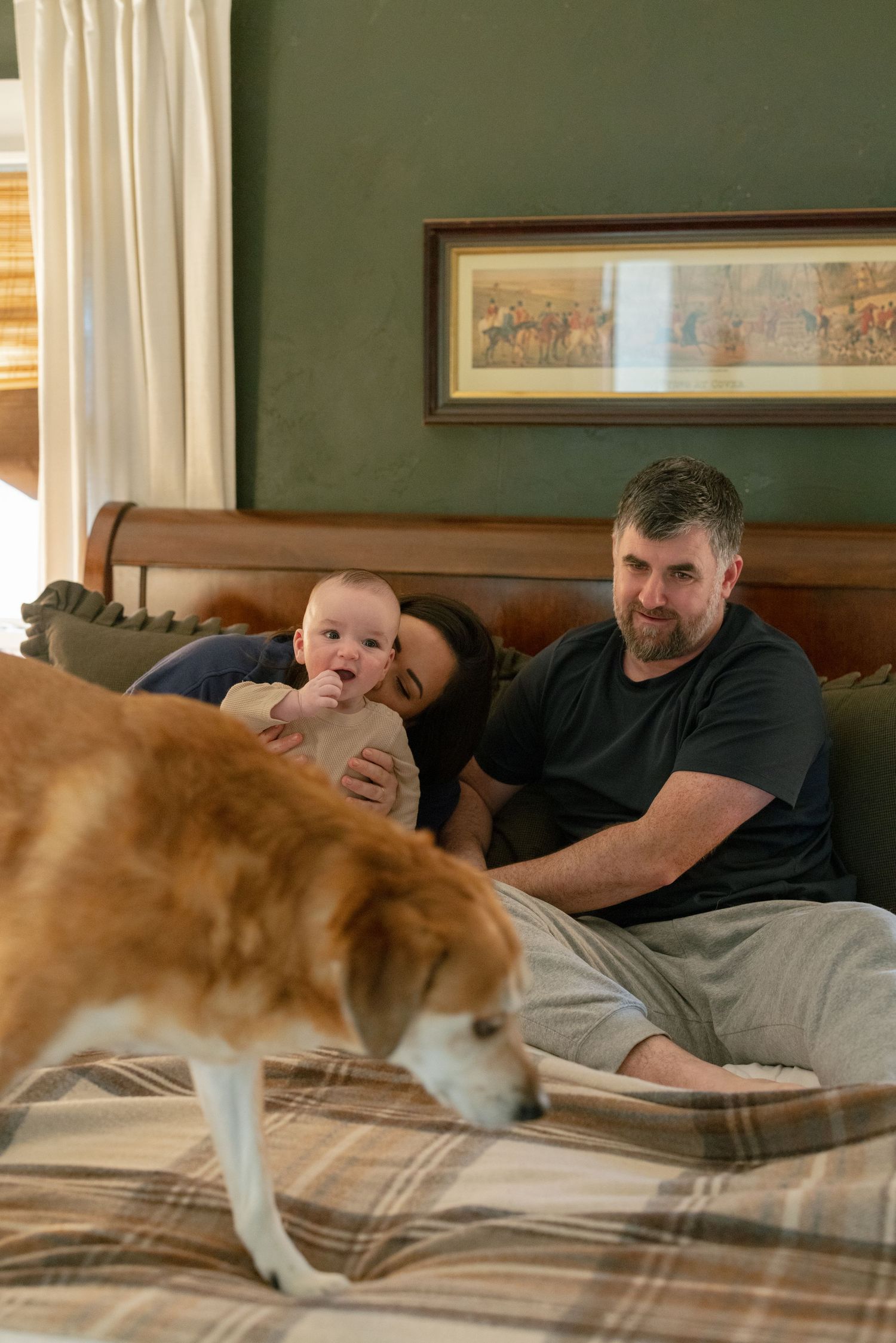 Family relaxes on bed with baby while brown and white dog explores plaid bedding in cozy bedroom setting.