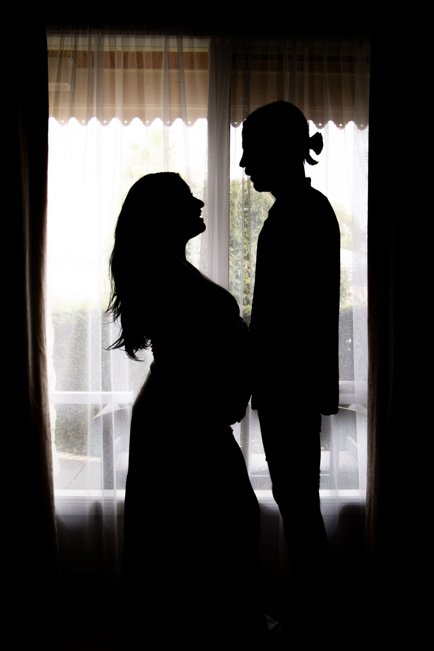 A silhouette of a couple stands by a window, the woman holding her pregnant belly as they face each other lovingly.