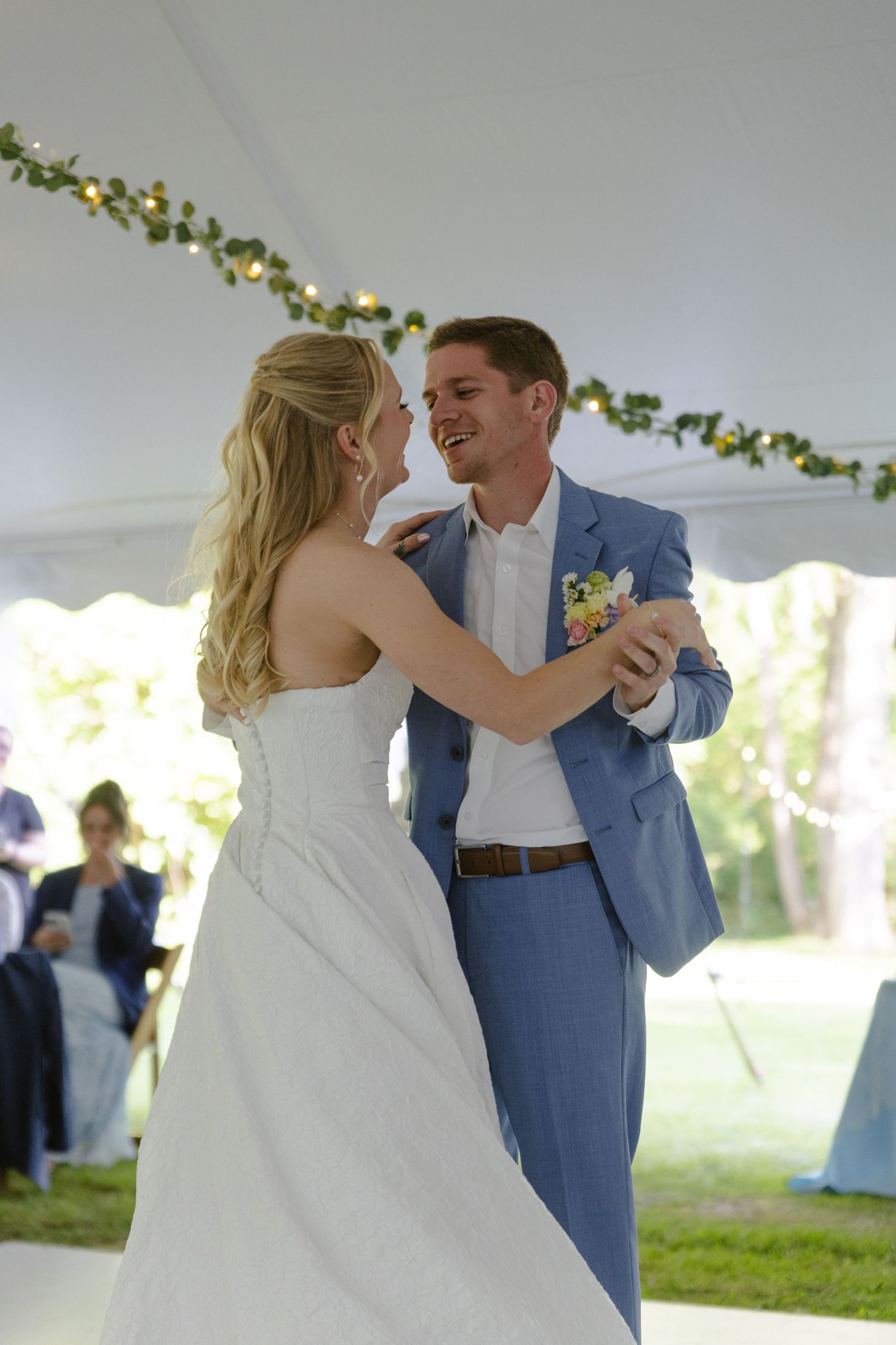 A couple shares their first dance under a tent decorated with greenery and string lights.