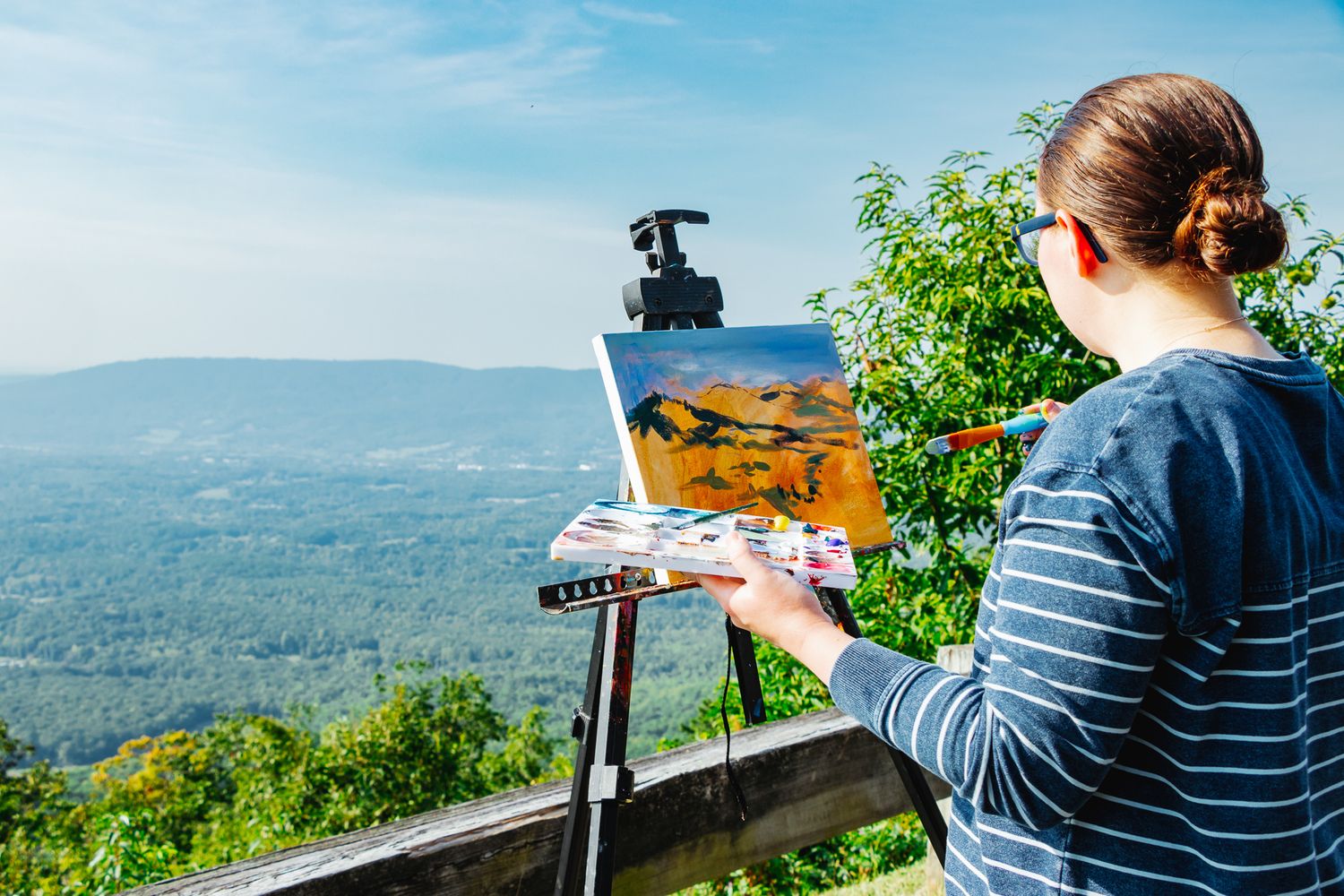 The back of an artist painting at an overlook, holding a palette and working on a canvas with wide mountain ranges below.