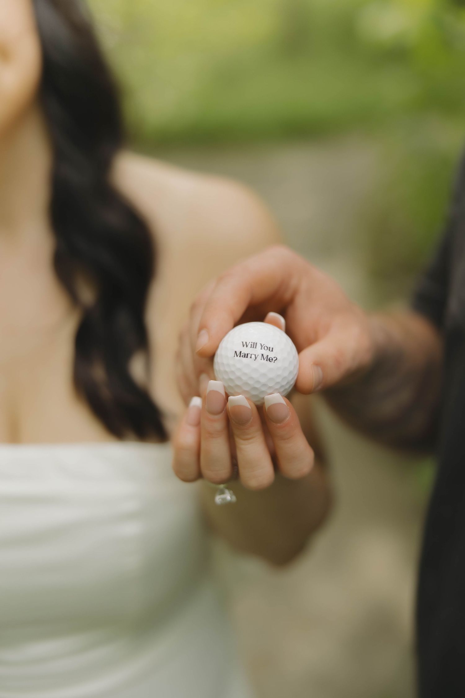 A white golf ball with 'Will You Marry Me?' text sits on a concrete surface with a thin line crack visible.