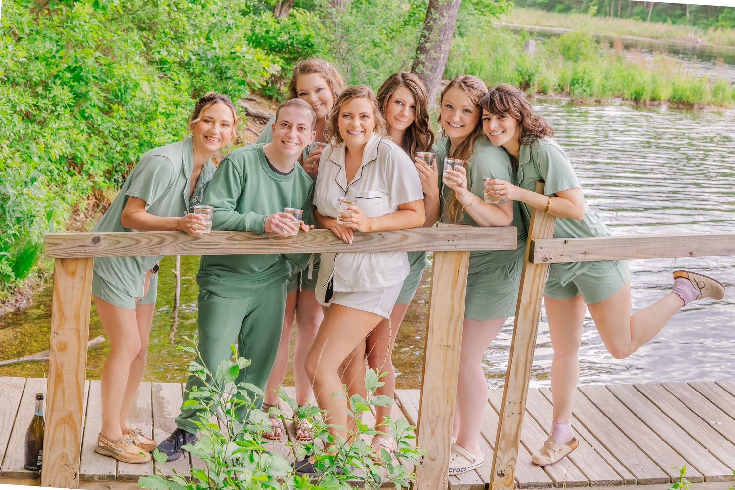 Group of people in matching sage green outfits pose together on wooden dock by lake on sunny summer day.