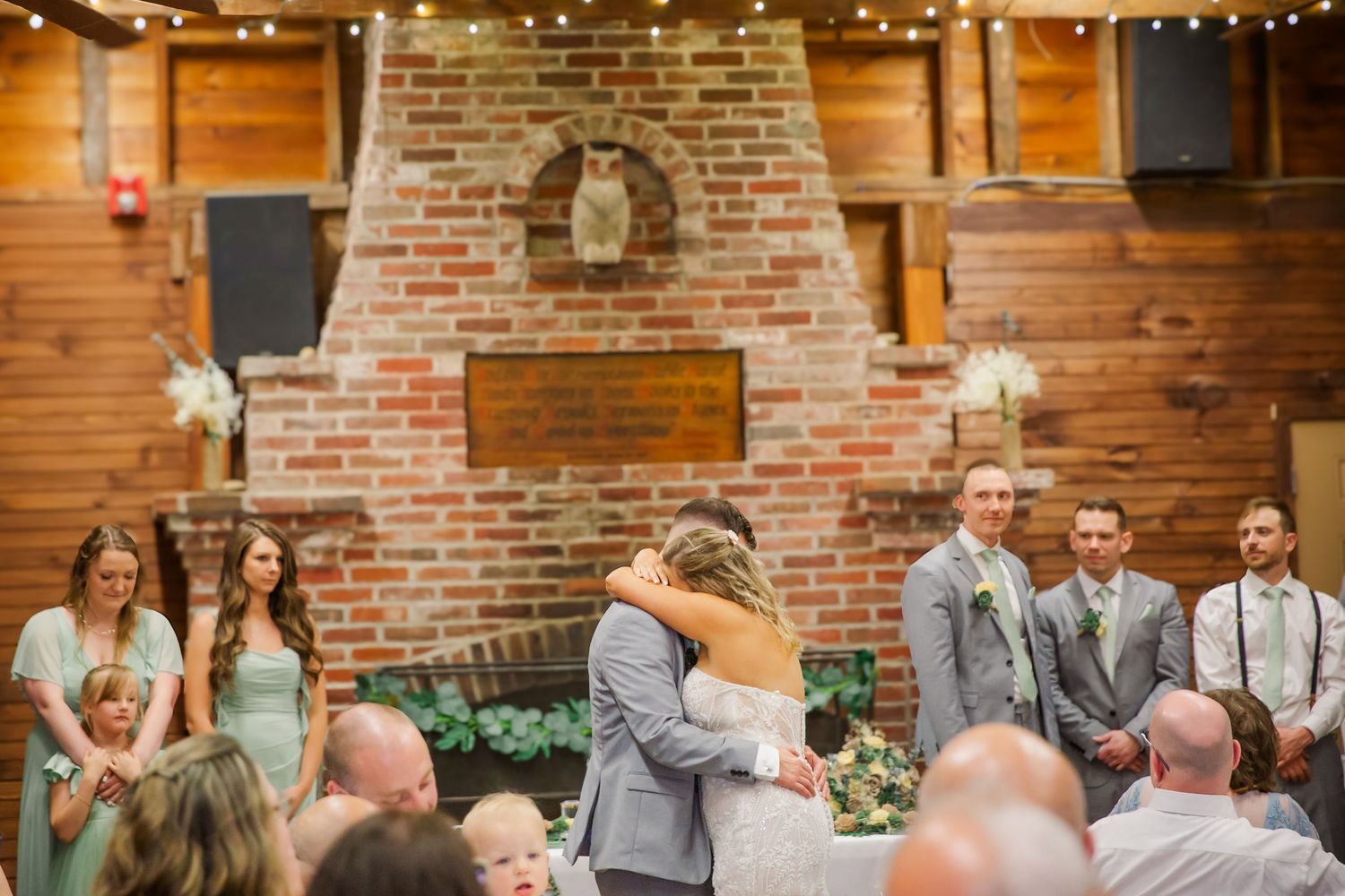 Wedding ceremony takes place in rustic venue with exposed brick fireplace and twinkling string lights overhead.