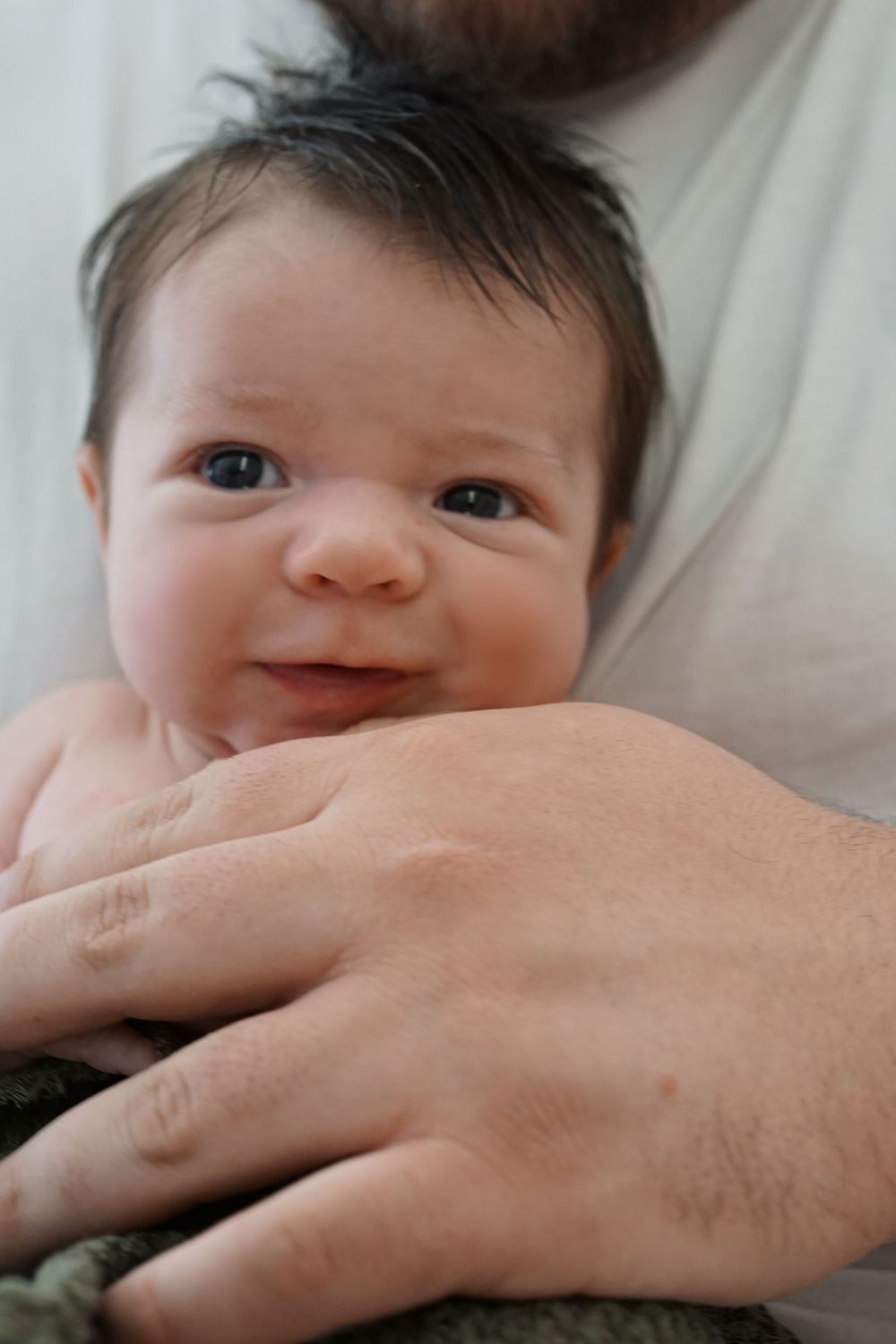 A baby smiles while lying down as an adult's hand rests gently near their face.