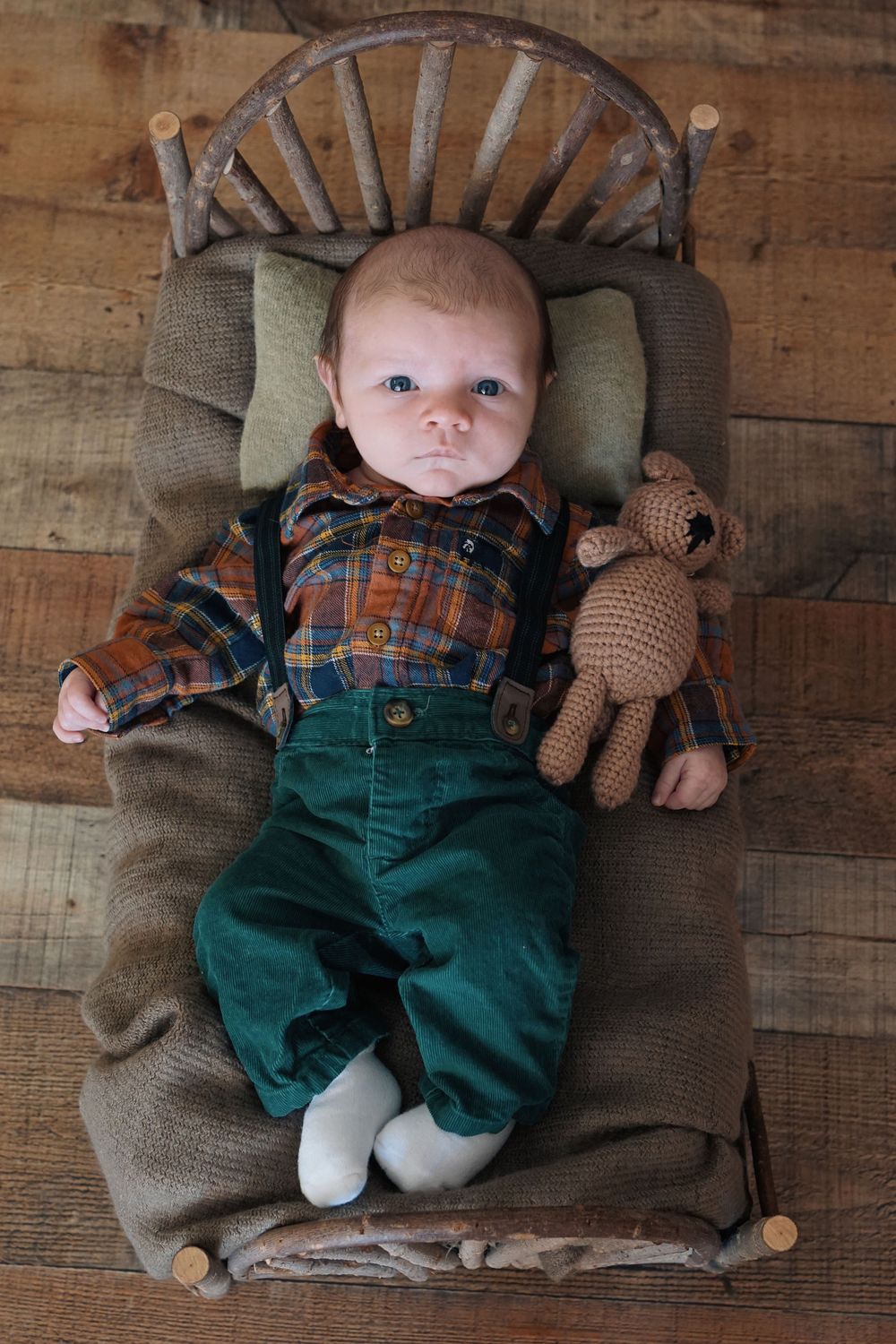 Small baby wearing plaid shirt and green overalls lies on rustic wooden bench with knitted teddy bear companion.