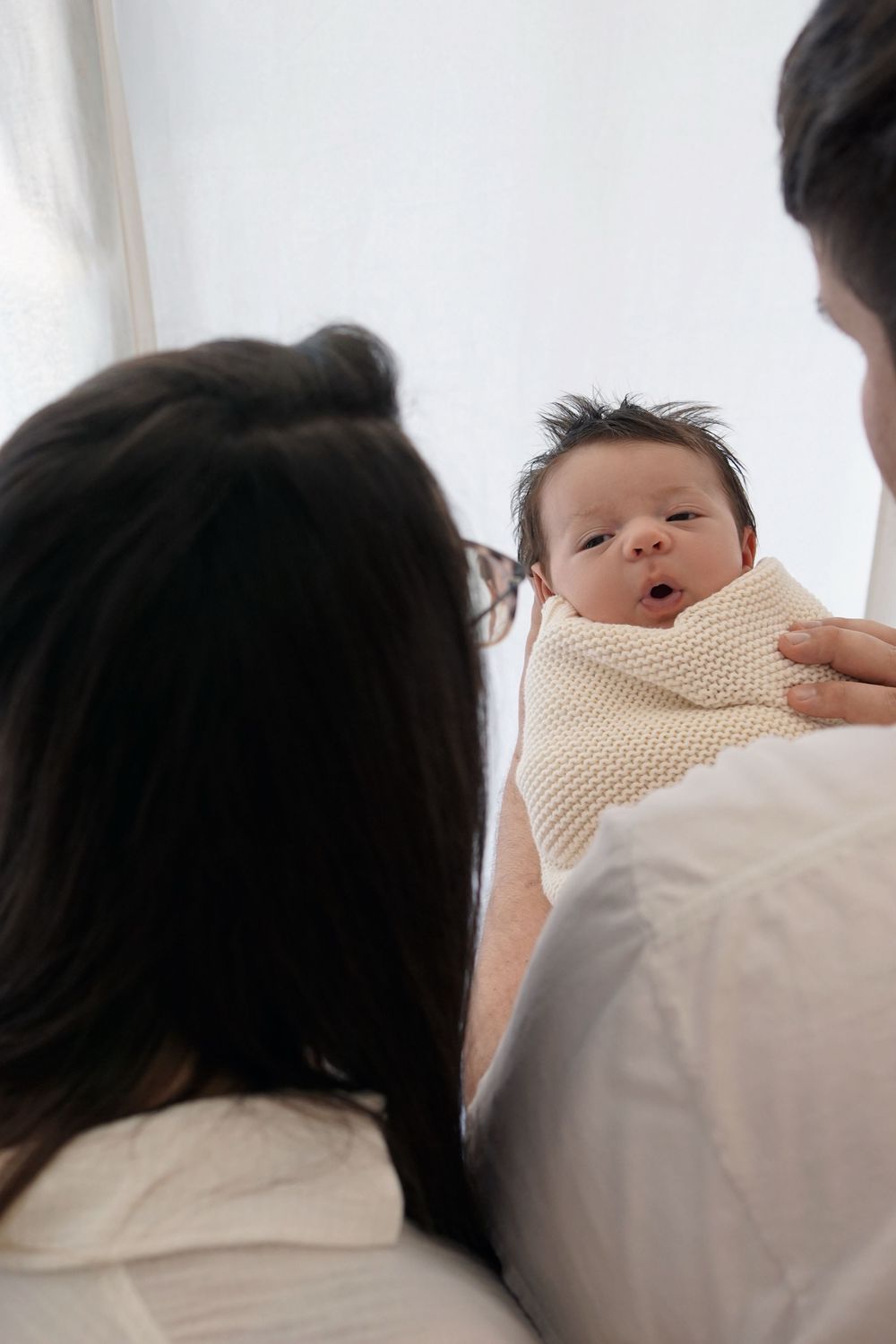A newborn baby swaddled in a cream-colored blanket is being held while making an adorable expression.