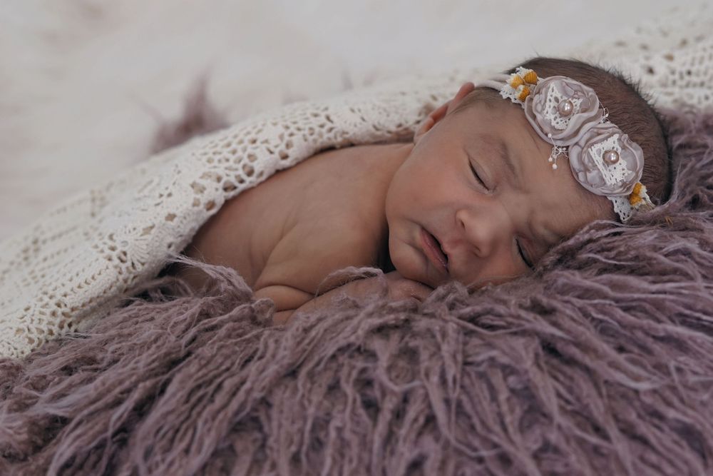 Sleeping newborn rests peacefully on purple textured blanket while wearing delicate floral headband.