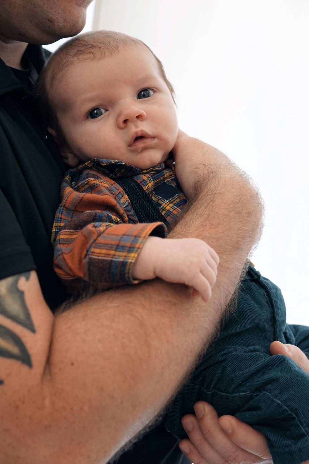 Baby in plaid shirt being held in tattooed arms against dark clothing in intimate portrait.