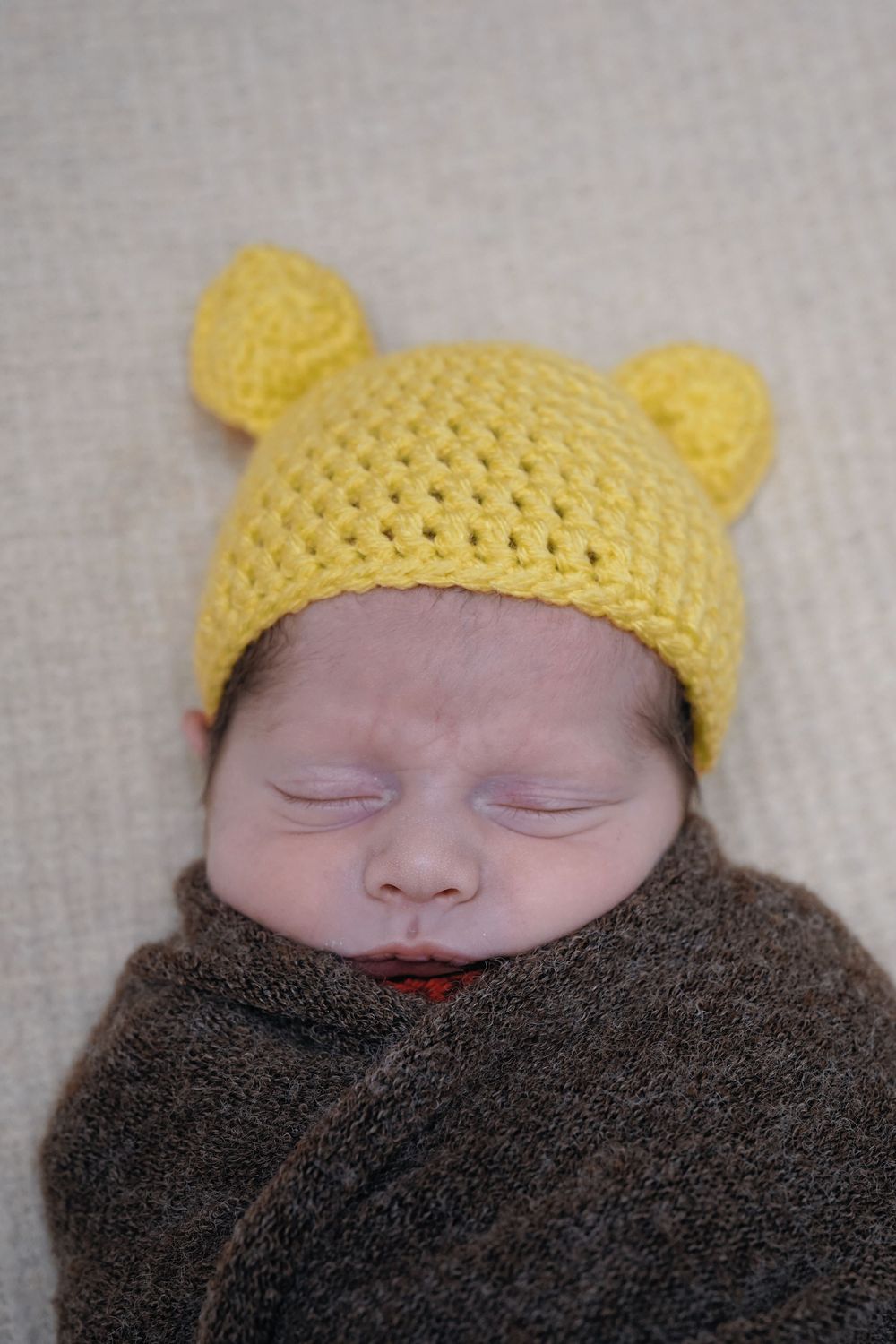 A sleeping baby wearing a yellow knitted hat with ears and wrapped in a brown blanket.