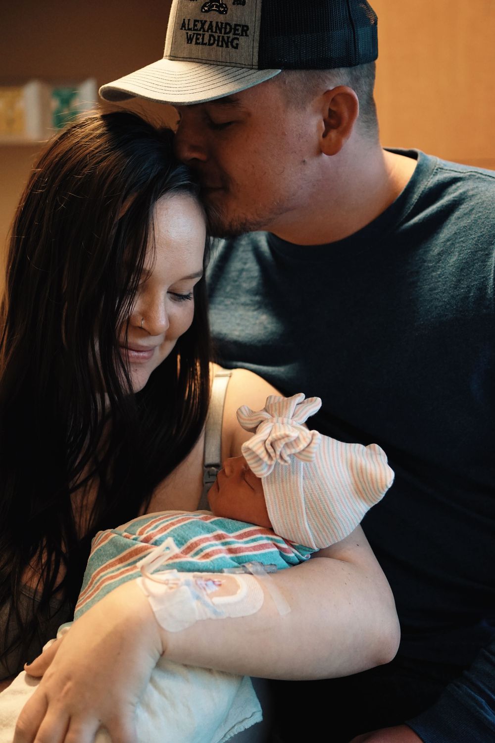 A couple shares an intimate moment while holding their newborn baby swaddled in white cloth during a tender family scene.