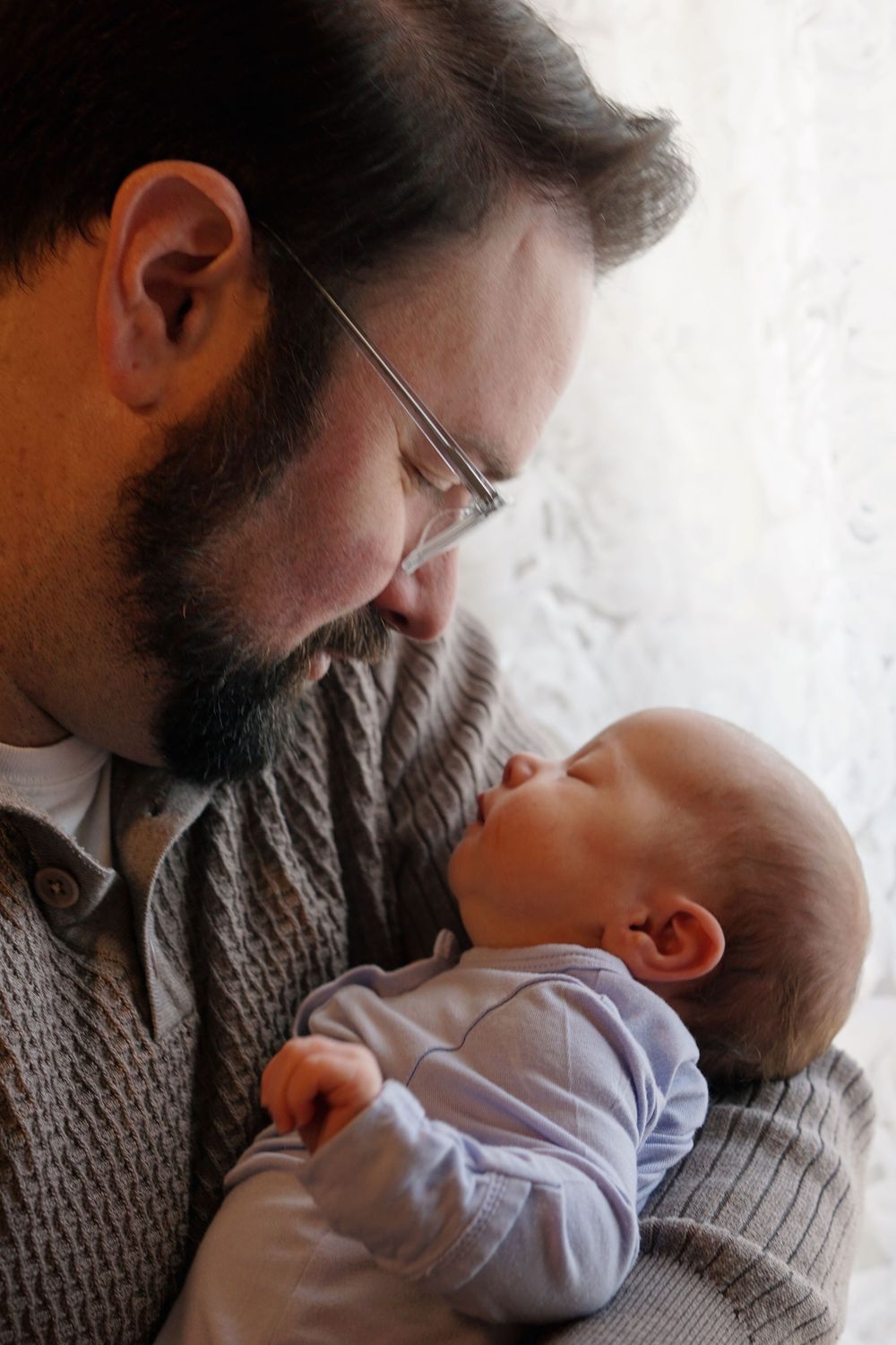 Close up of a bearded person wearing glasses and a gray sweater tenderly holding a small baby in a light blue onesie.