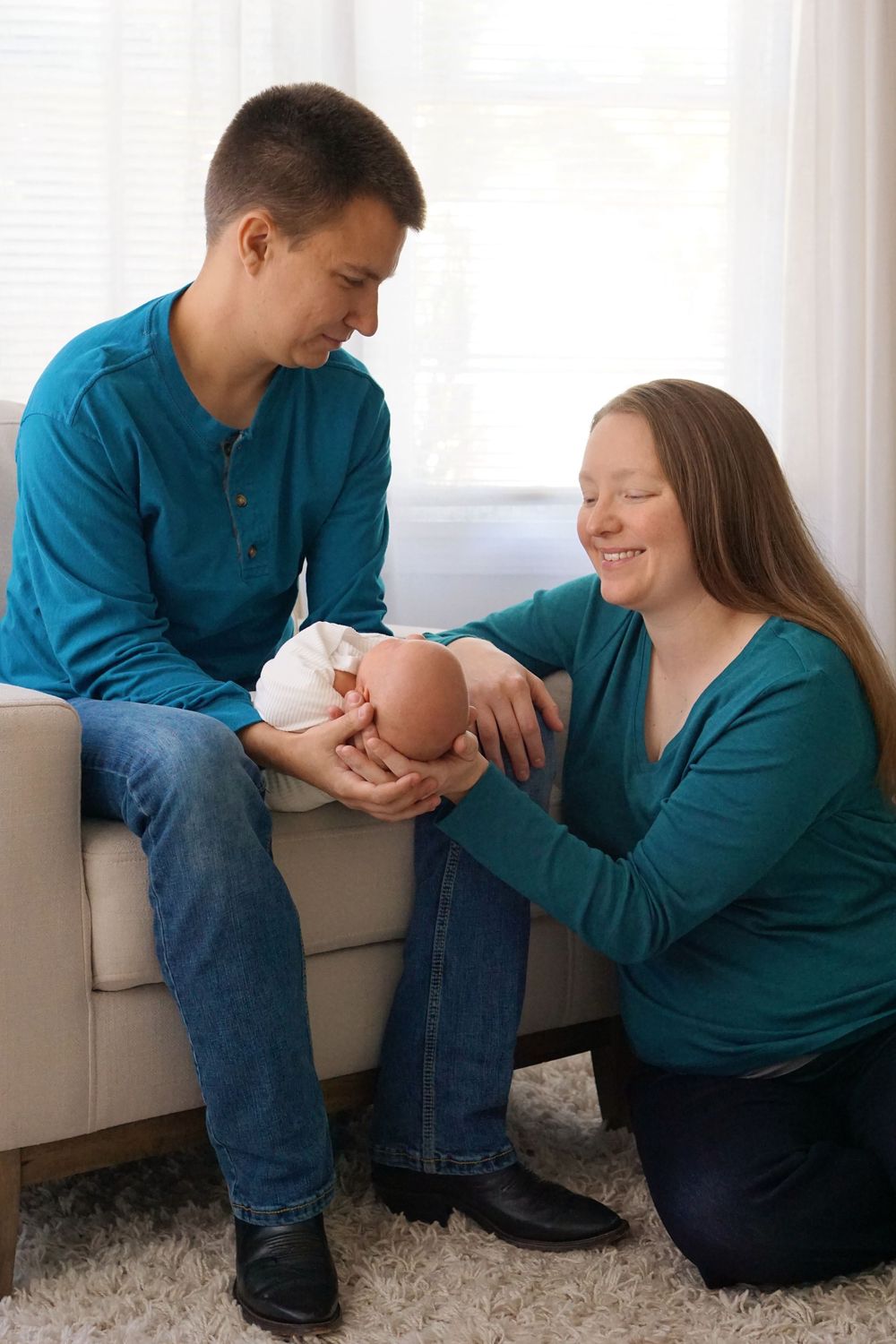 A couple sits together on a couch sharing an intimate moment with their newborn baby in their bright living room.