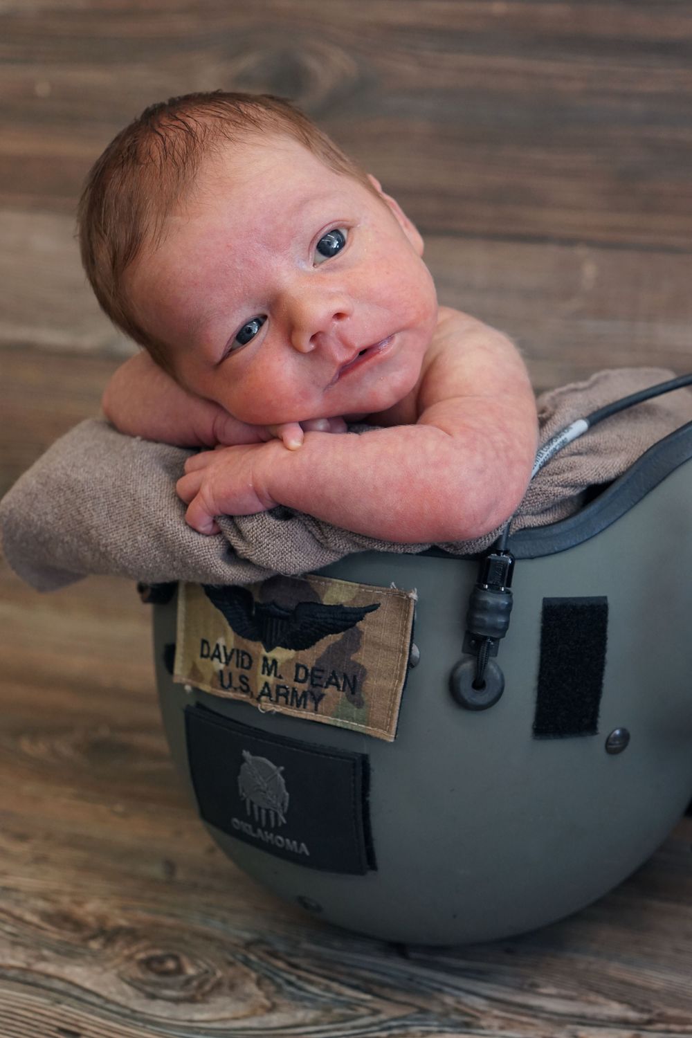 A baby rests their head on military helmet with woodland background.