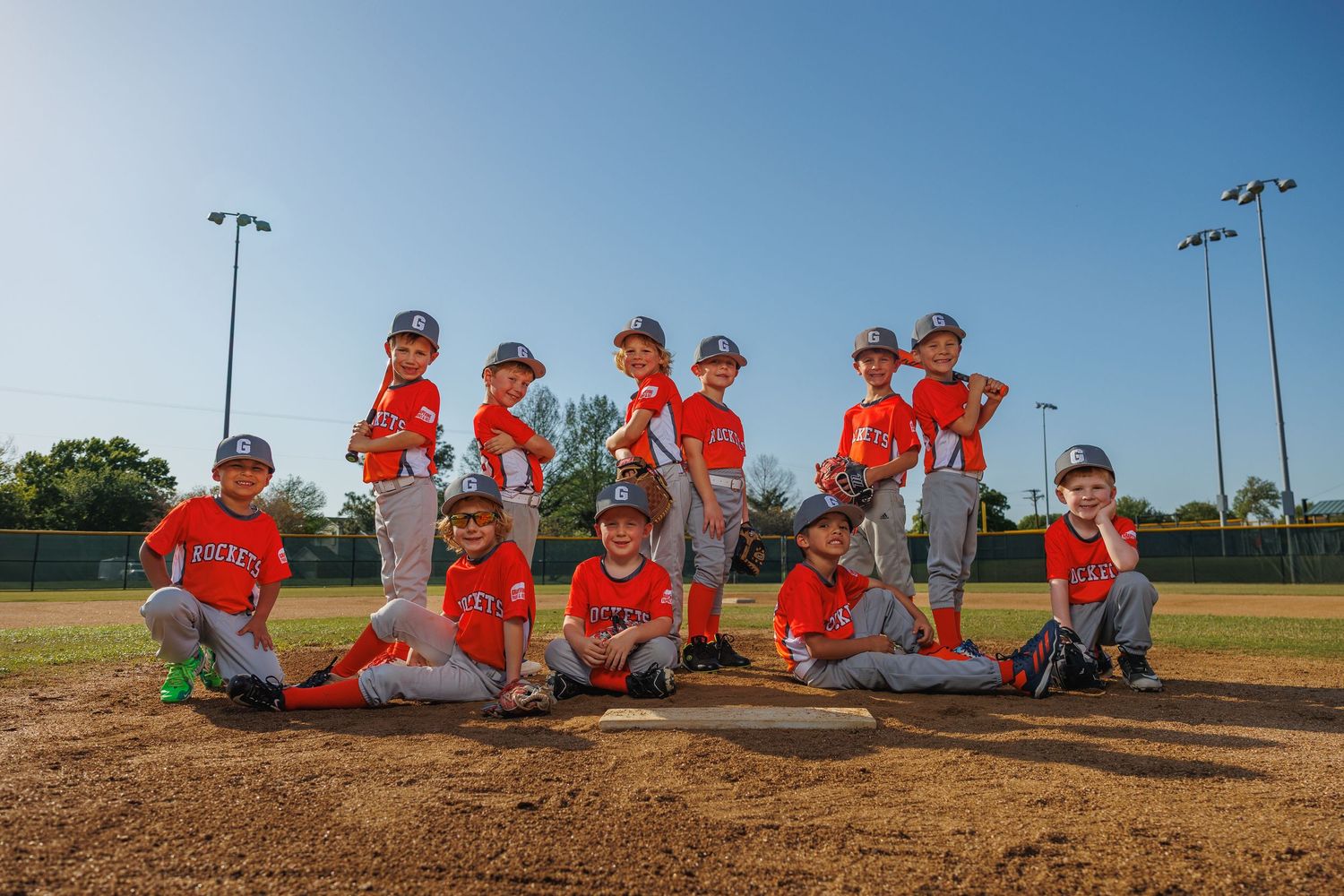 Youth baseball team poses playfully on pitcher's mound wearing matching orange jerseys at sunset on baseball field.