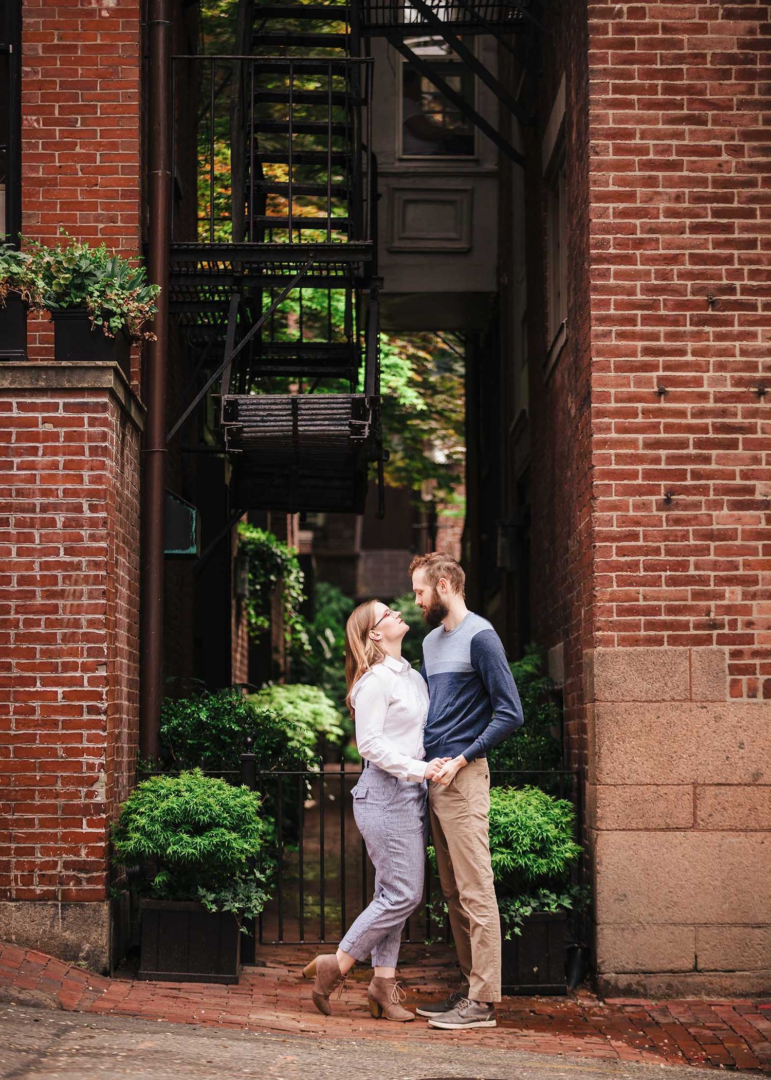 A romantic portrait of a couple holding hands and smiling at each other in a narrow brick alleyway in Beacon Hill with ivy-draped walls