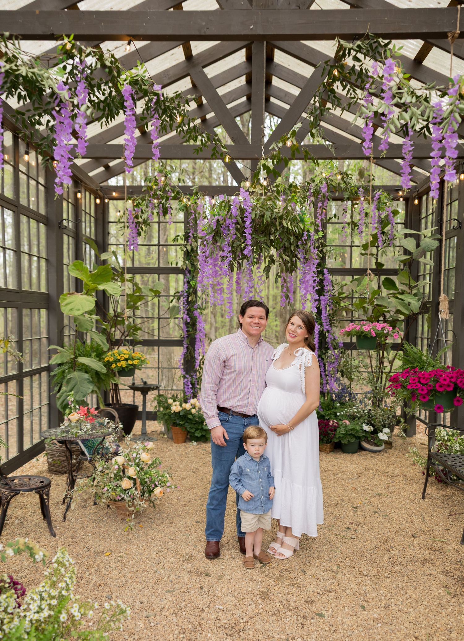 Family poses together inside a greenhouse decorated with hanging purple wisteria flowers and potted plants.