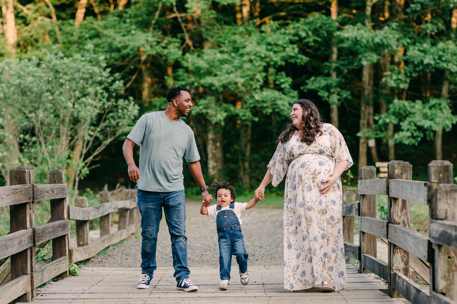 Family walking together on a wooden bridge path through a lush forest during a maternity shoot at Southford Falls, CT