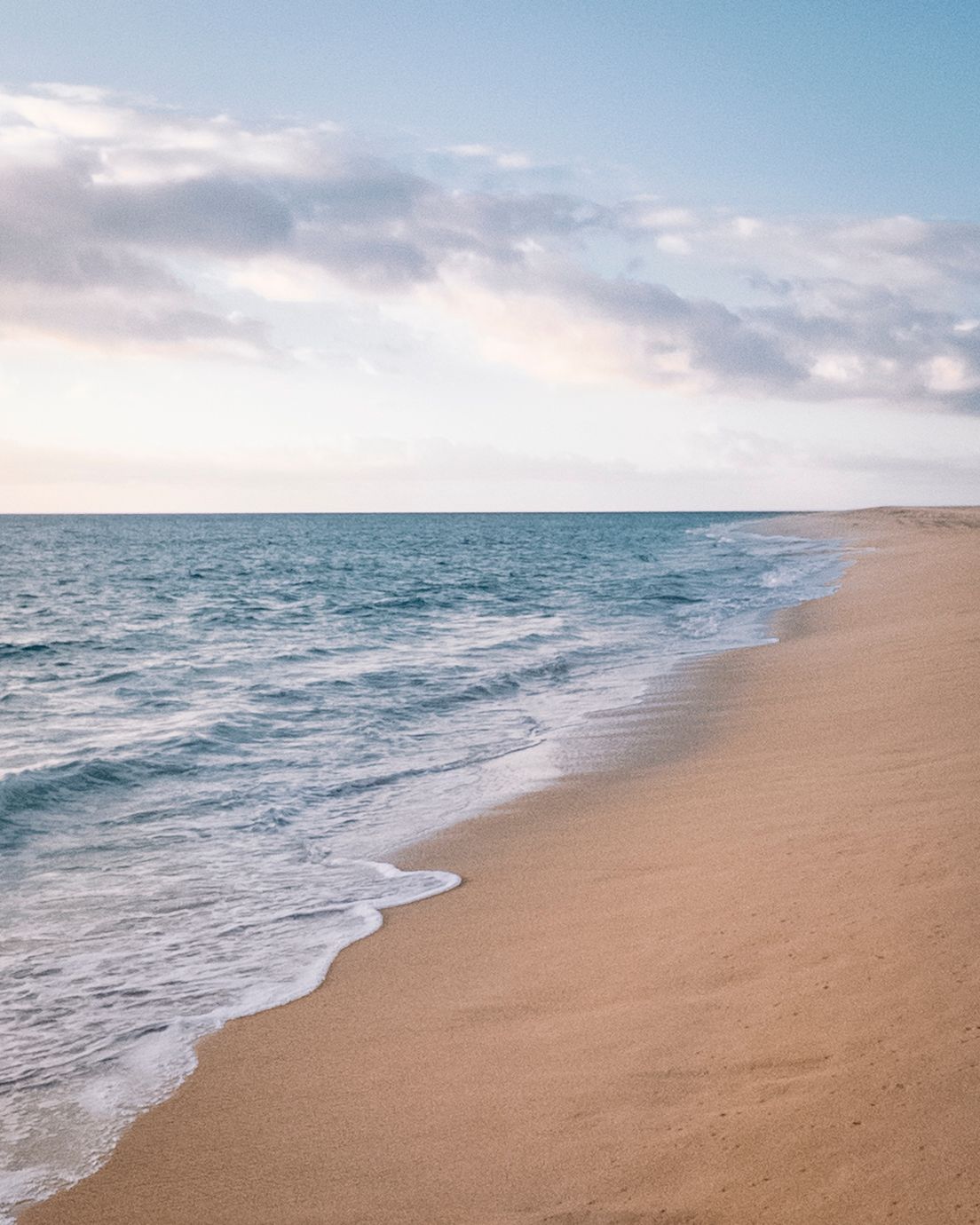 Serene wide sandy beach with calm turquoise ocean waves under a pastel cloudy sky.