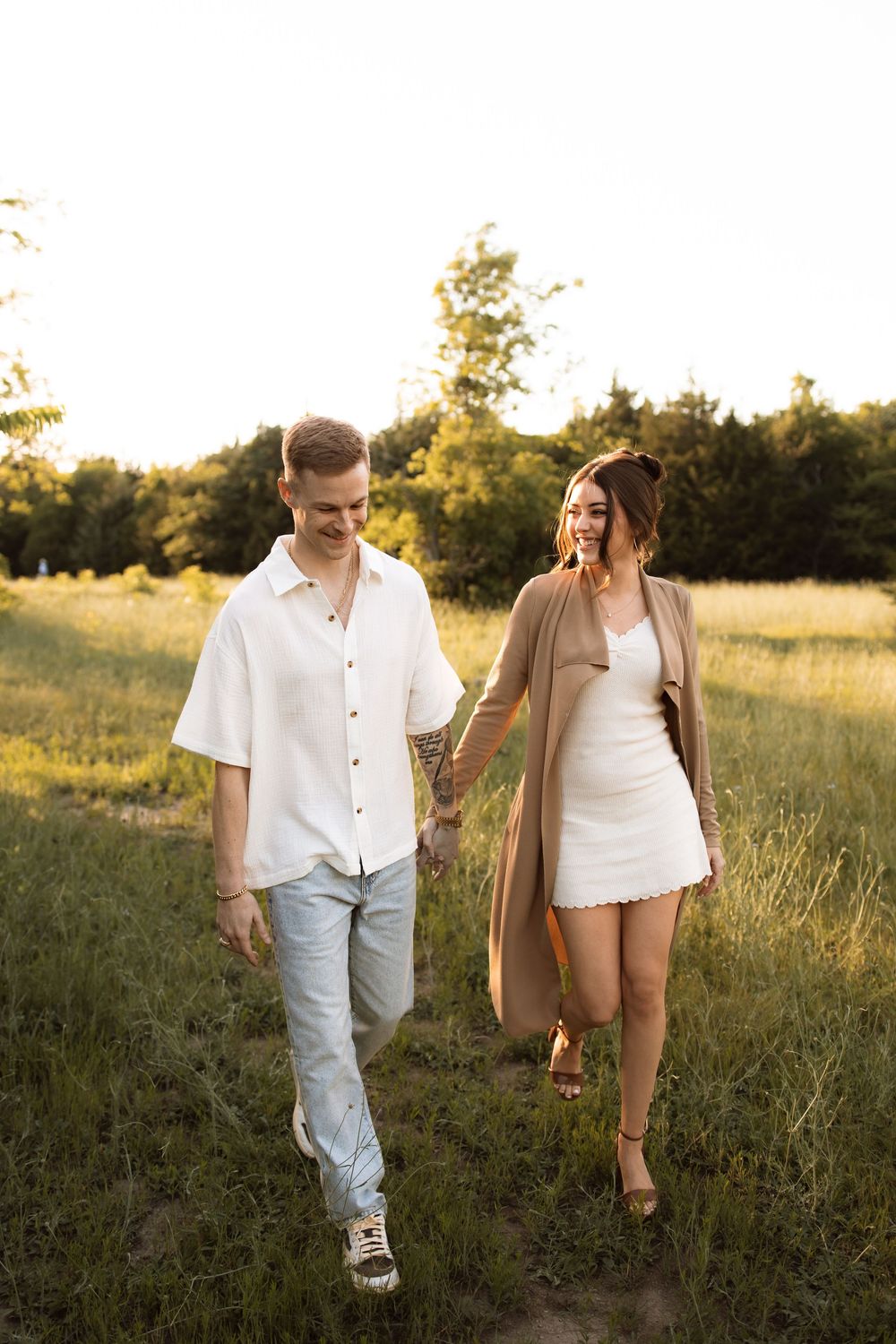 A couple strolls together through a sun-drenched meadow wearing casual white and denim outfits at golden hour.