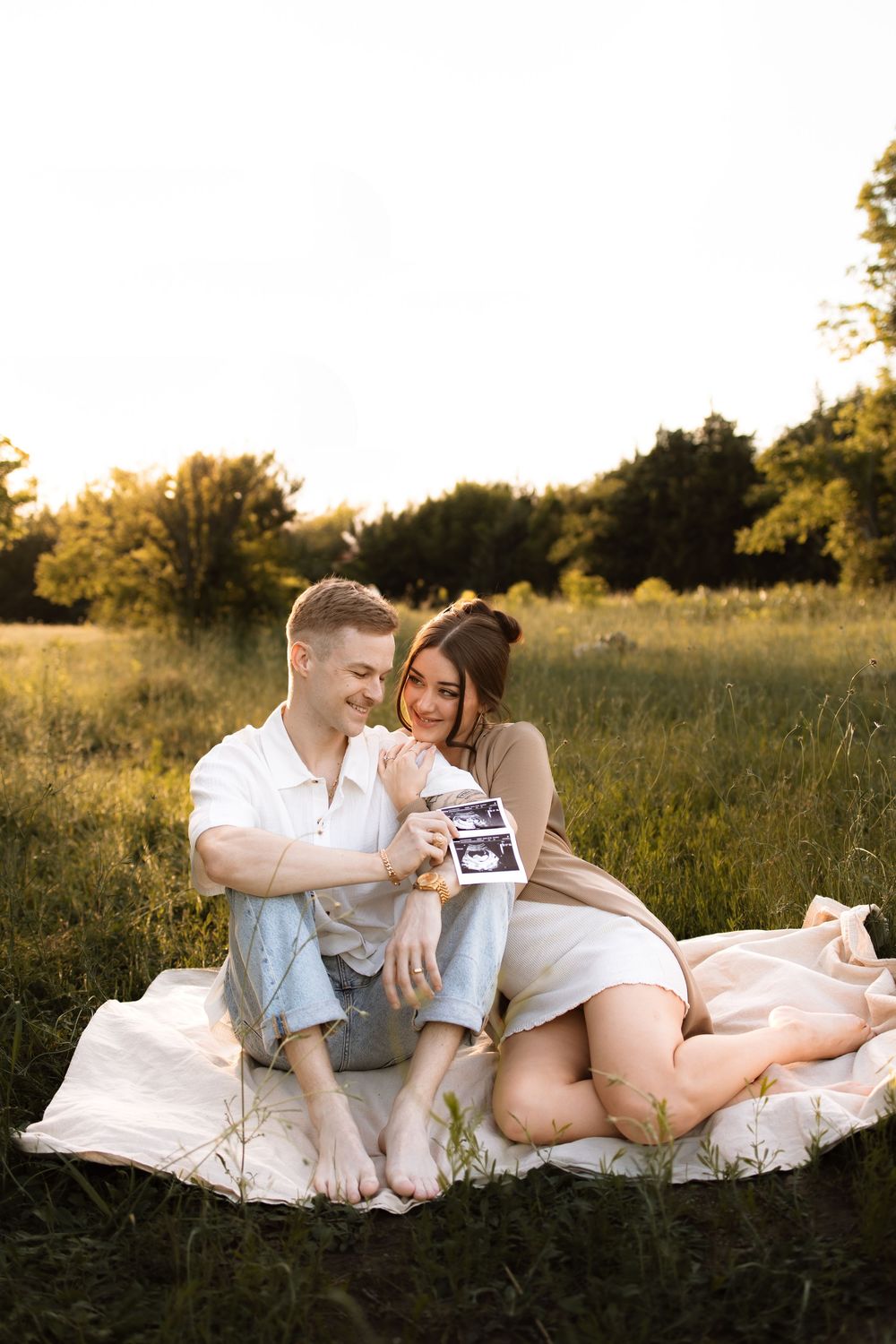 Expecting couple relaxes on white blanket in sunlit field while looking at ultrasound photos together.