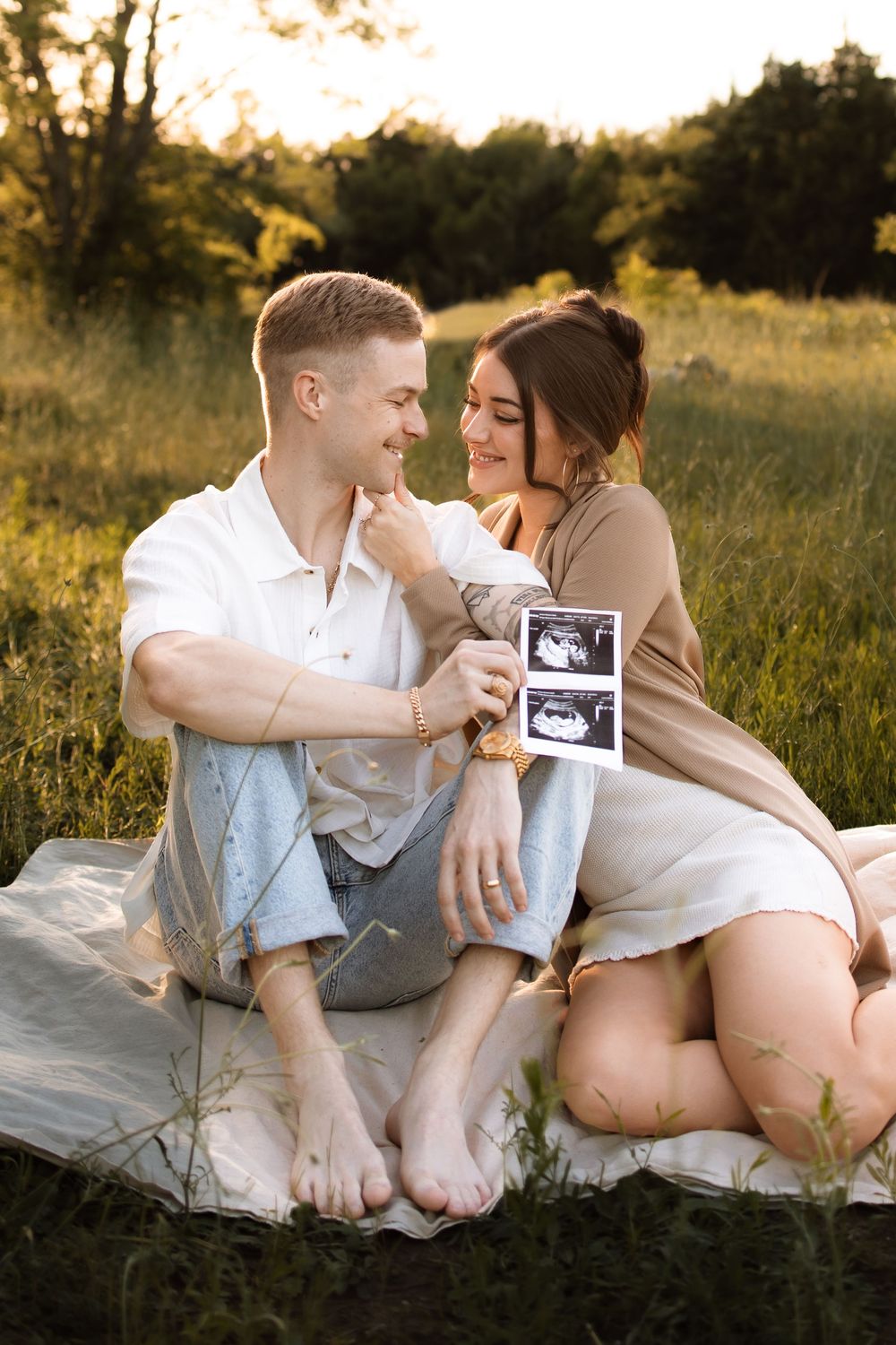 A couple sits on a blanket in a field sharing a tender moment while holding ultrasound photos.