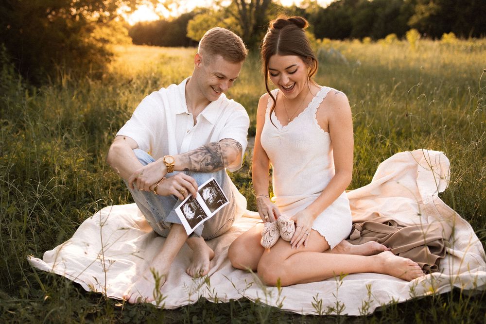 A couple looks lovingly at ultrasound photos while sitting on a blanket in a sunny field.