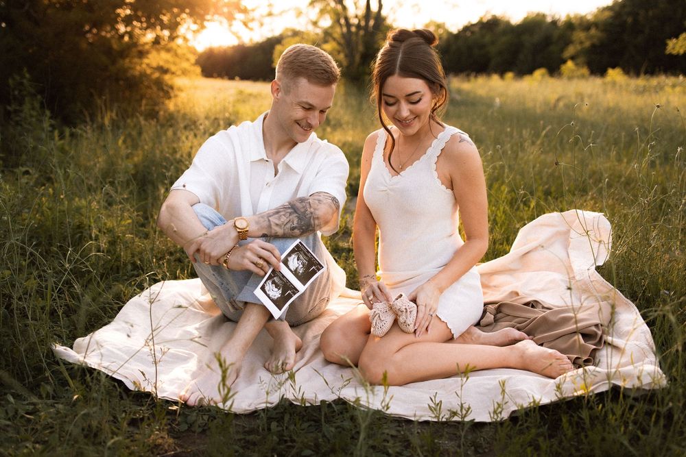 Expecting couple sits on blanket in field looking at ultrasound photos during sunset.