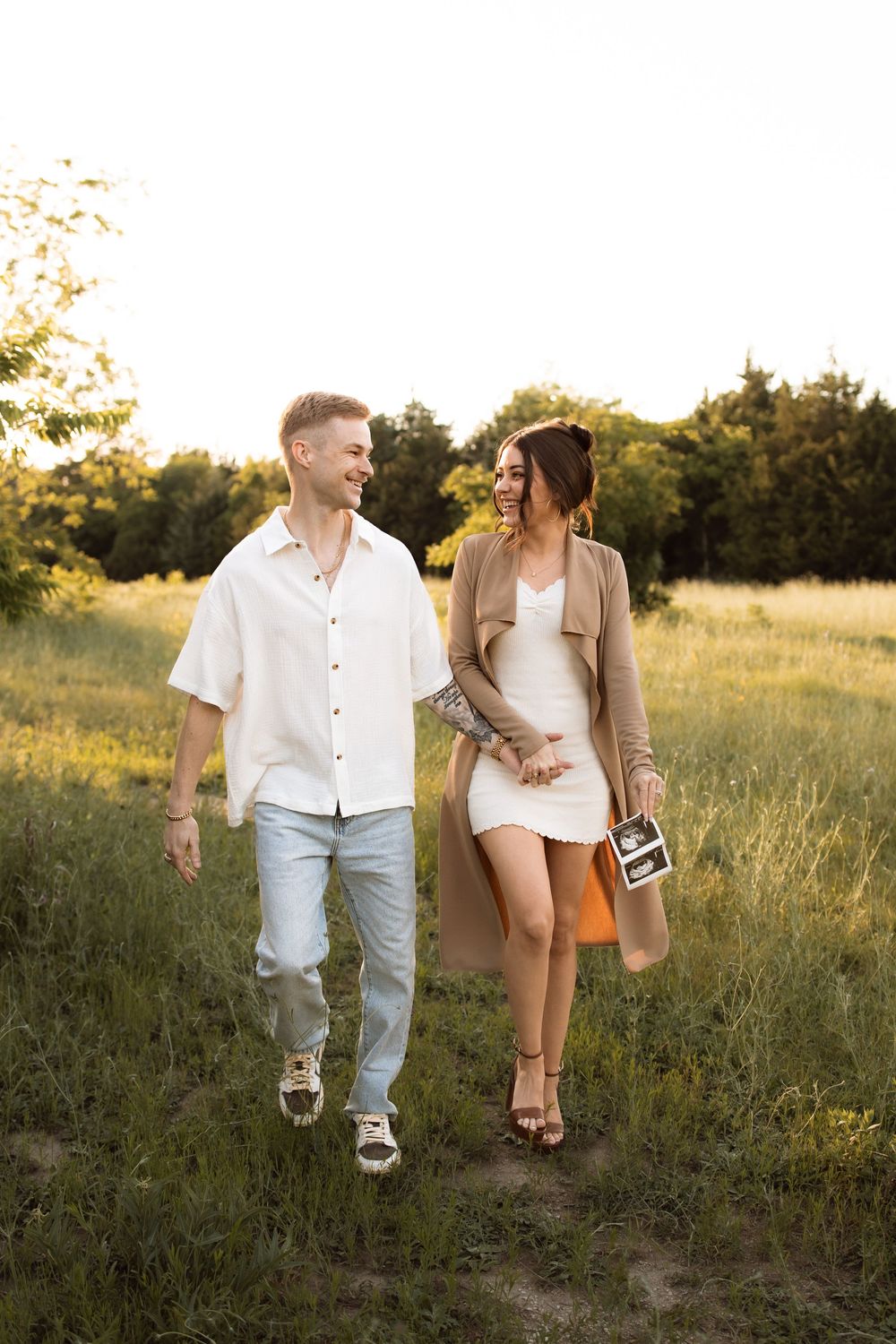 A couple walks together in a golden-lit field during sunset while holding ultrasound photos.