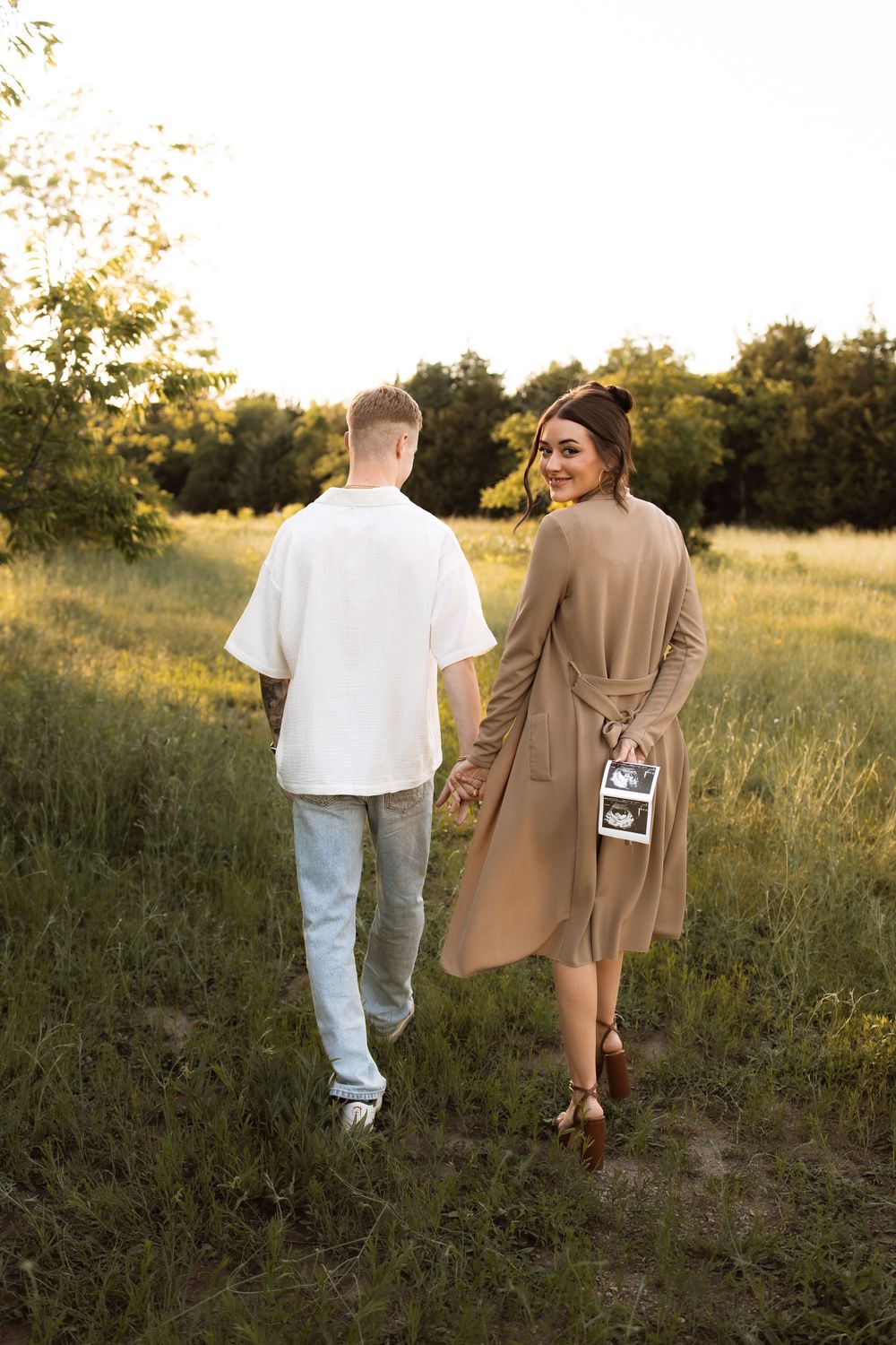 Couple walking in a golden field at sunset, with her wearing a tan coat and holding ultrasound photos.