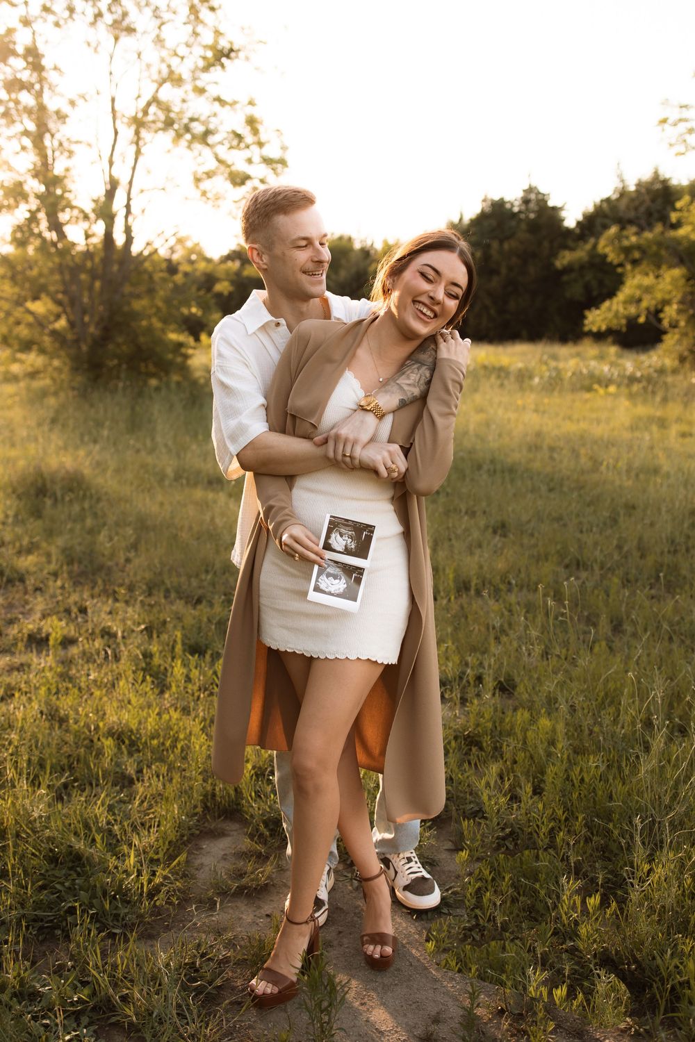 Joyful couple embracing in a sunlit meadow while holding ultrasound pictures and sharing a happy moment.