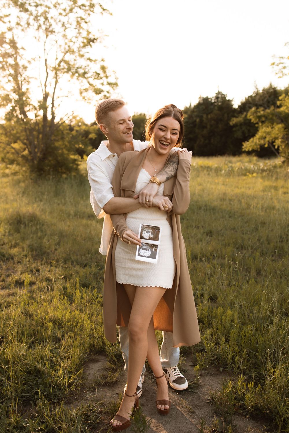 A couple share an intimate moment outdoors at sunset while holding ultrasound photos.