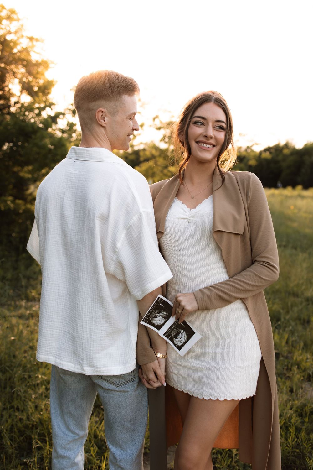 Happy couple in casual attire standing close together in a field holding ultrasound photos at golden hour.