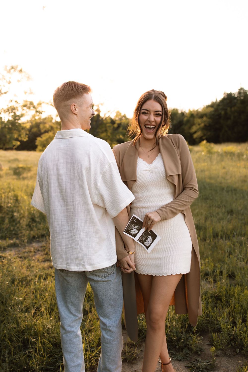 A couple shares a joyful moment while holding ultrasound photos in a summer field at golden hour.