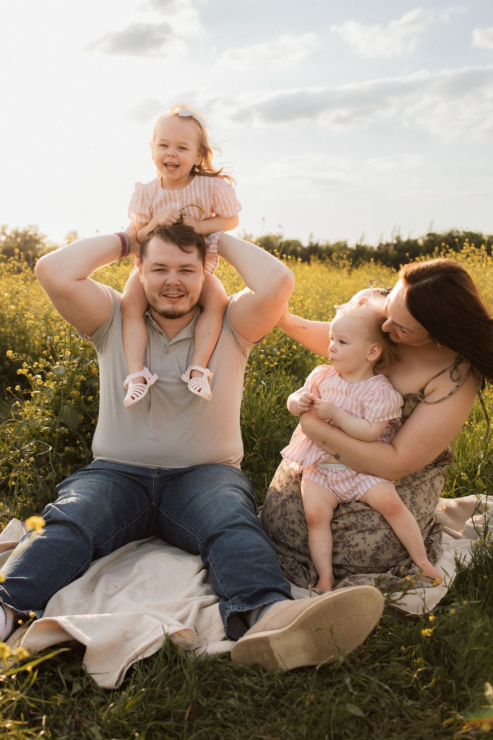 Family playing and laughing together on picnic blanket surrounded by yellow flowers at sunset.
