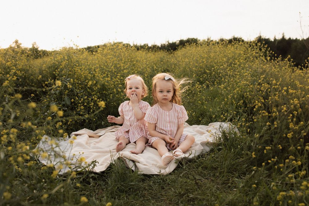 Two children in pink dresses sit together on white blanket amongst a field of yellow wildflowers.