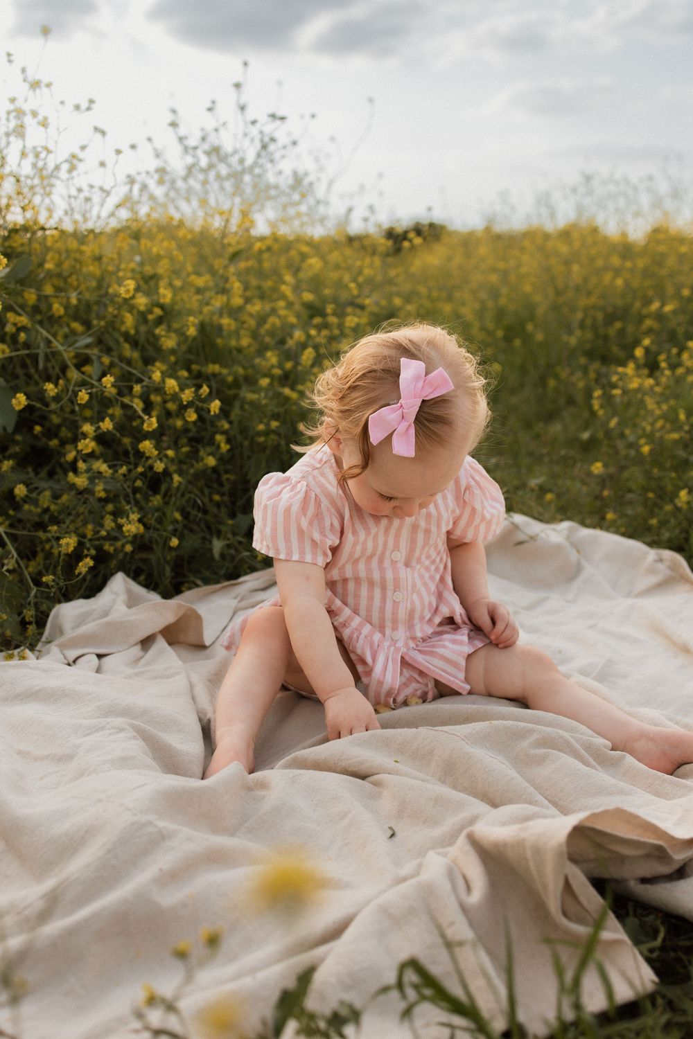 Child in pink striped dress sitting on blanket in field of yellow wildflowers.