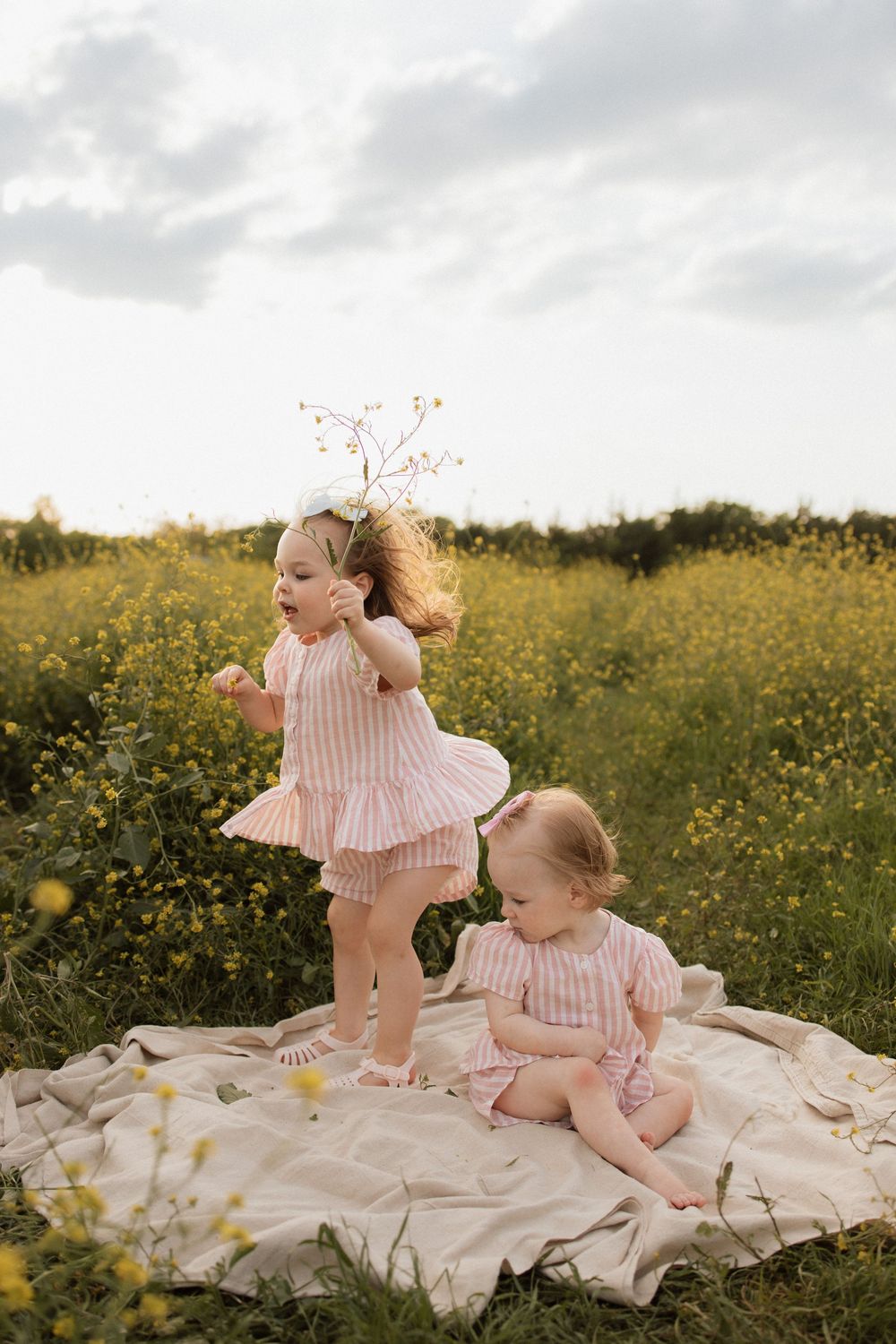 Two children in matching pink striped outfits play on a blanket in a field of yellow wildflowers.