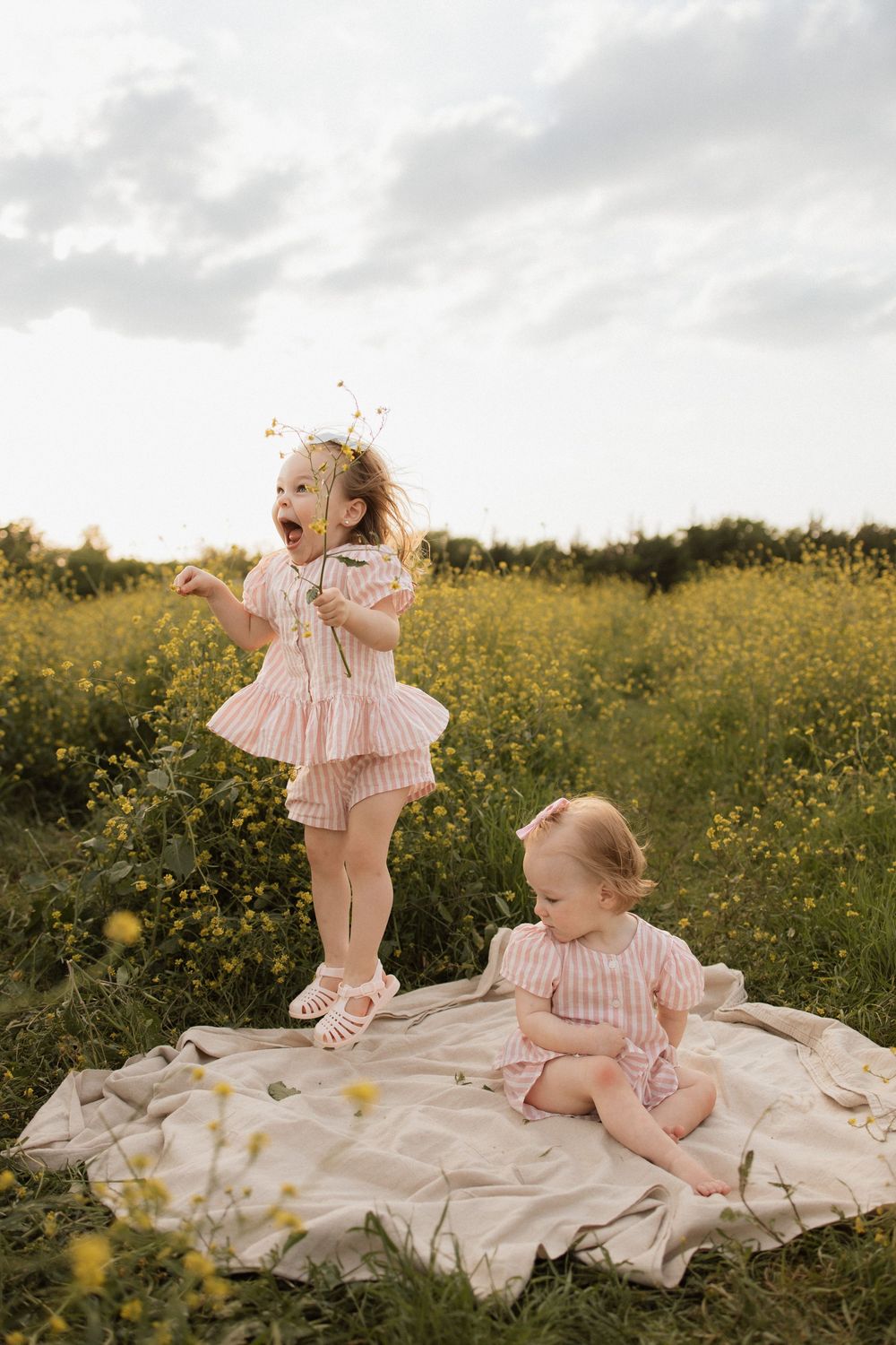Two small children in pink striped outfits play on a blanket in a wildflower meadow at sunset.