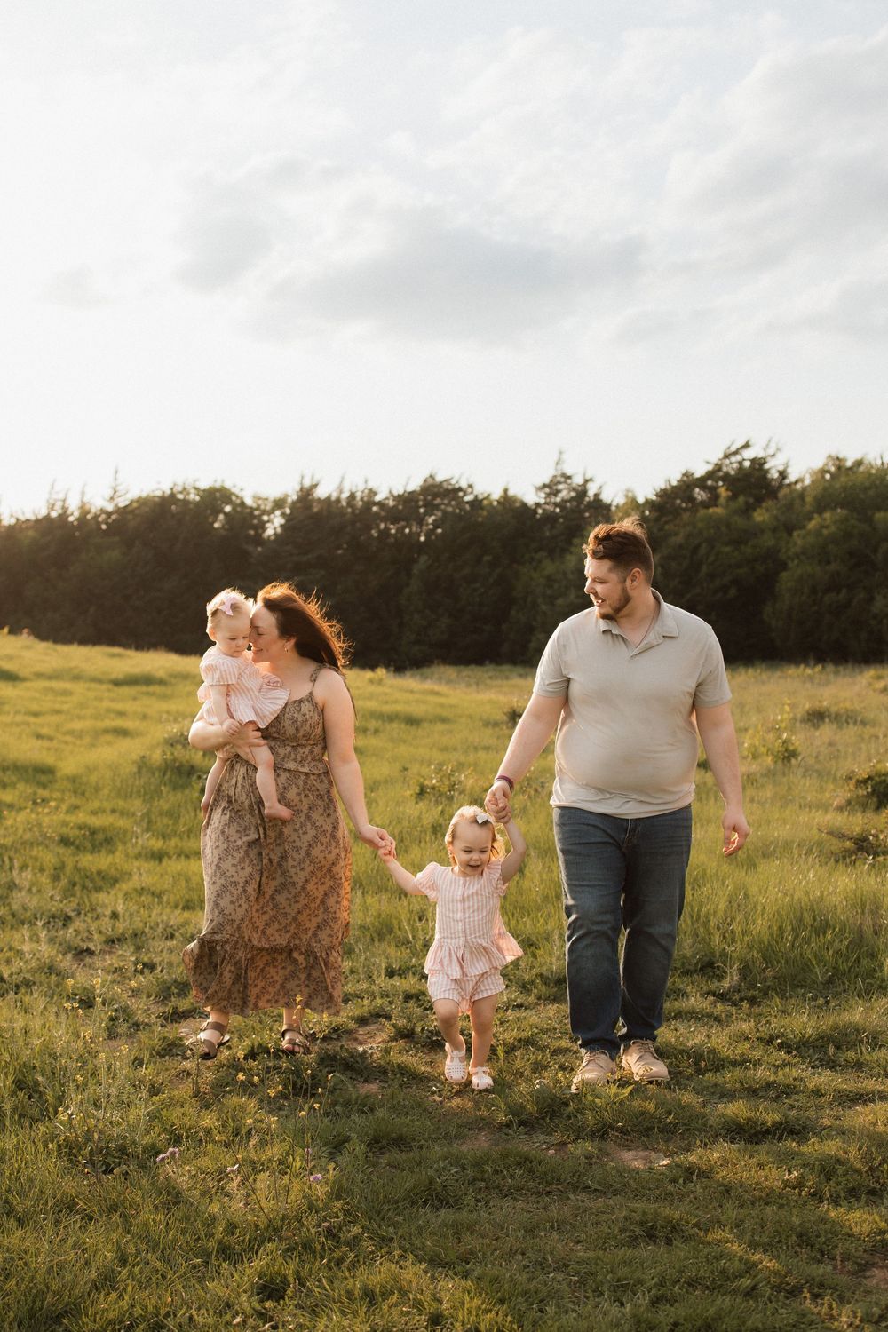 A family walks together through a sunlit meadow at golden hour with trees in the background.