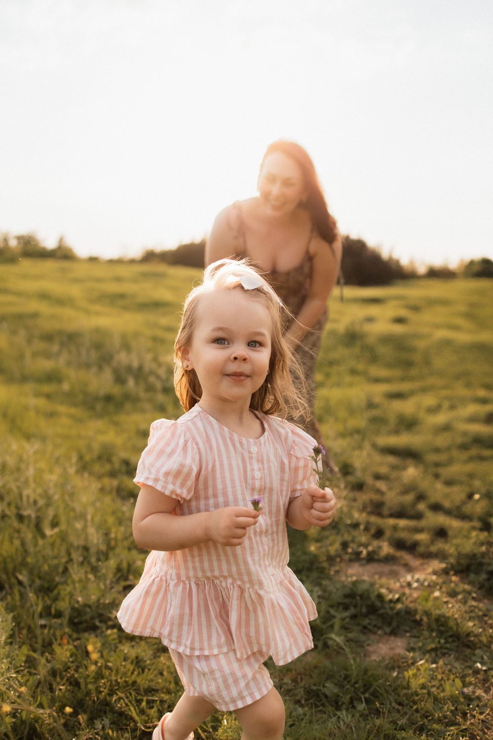 A toddler in a pink striped dress runs through a sunlit meadow during golden hour.