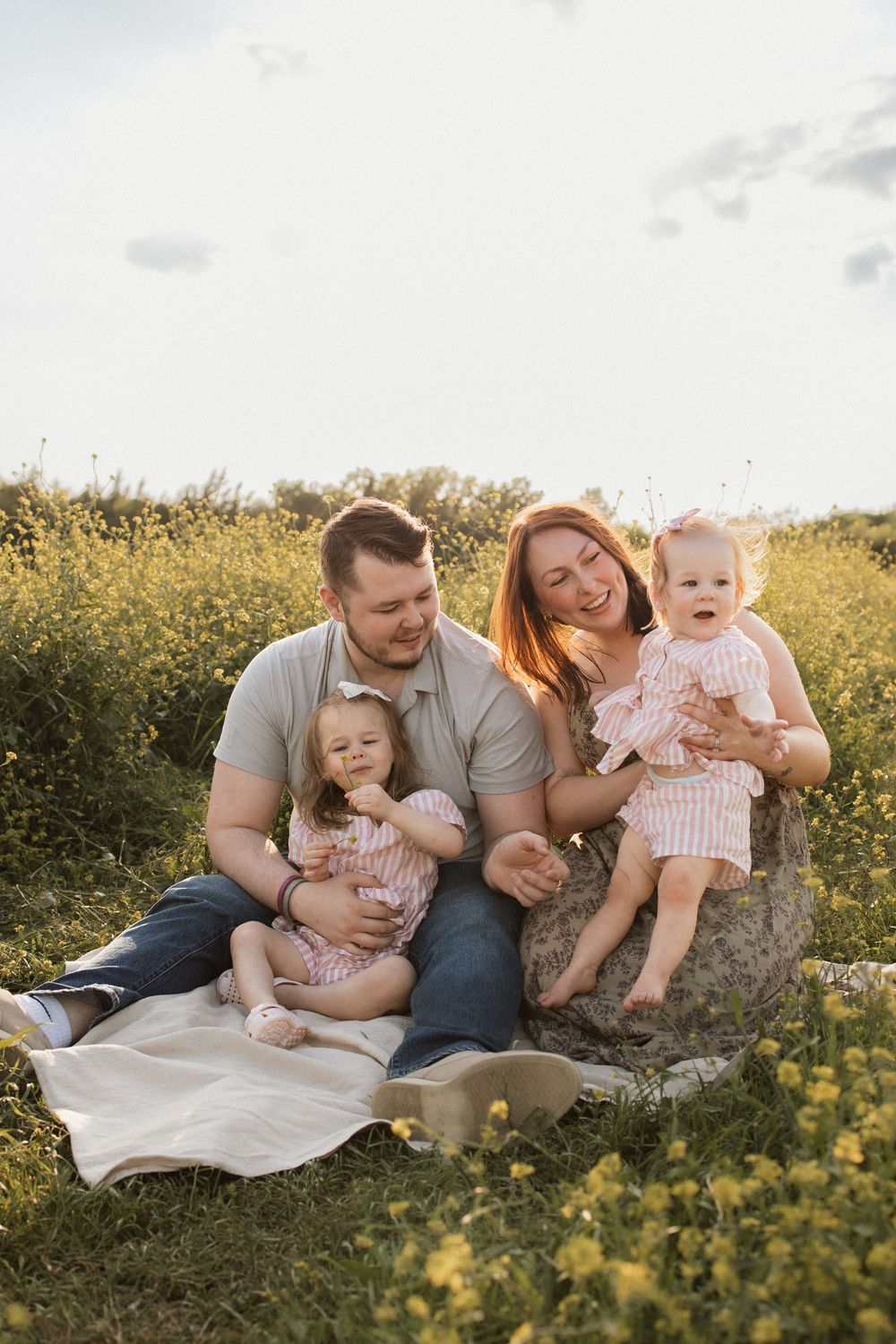 Family sitting on blanket in field of yellow wildflowers during golden hour sunset.