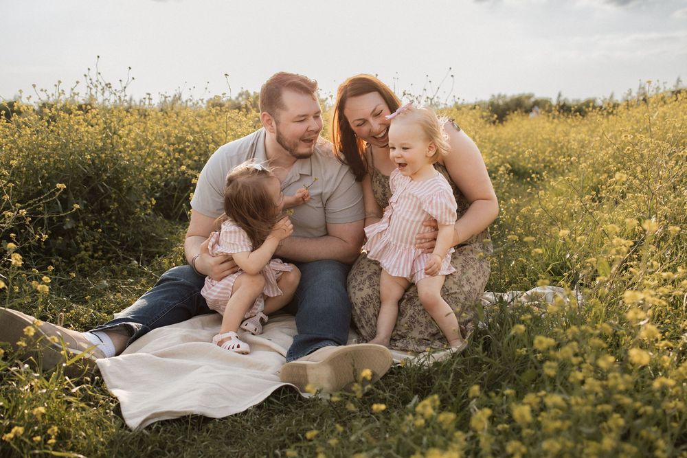Family sits together on blanket in blooming yellow wildflower field during golden hour sunset.