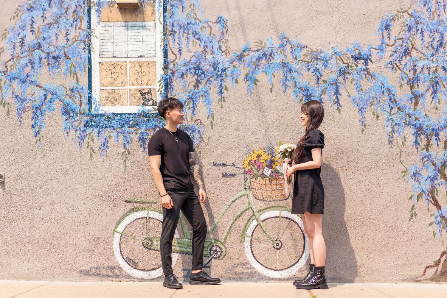 Couple standing by vintage bicycle against pink wall with climbing blue wisteria flowers.