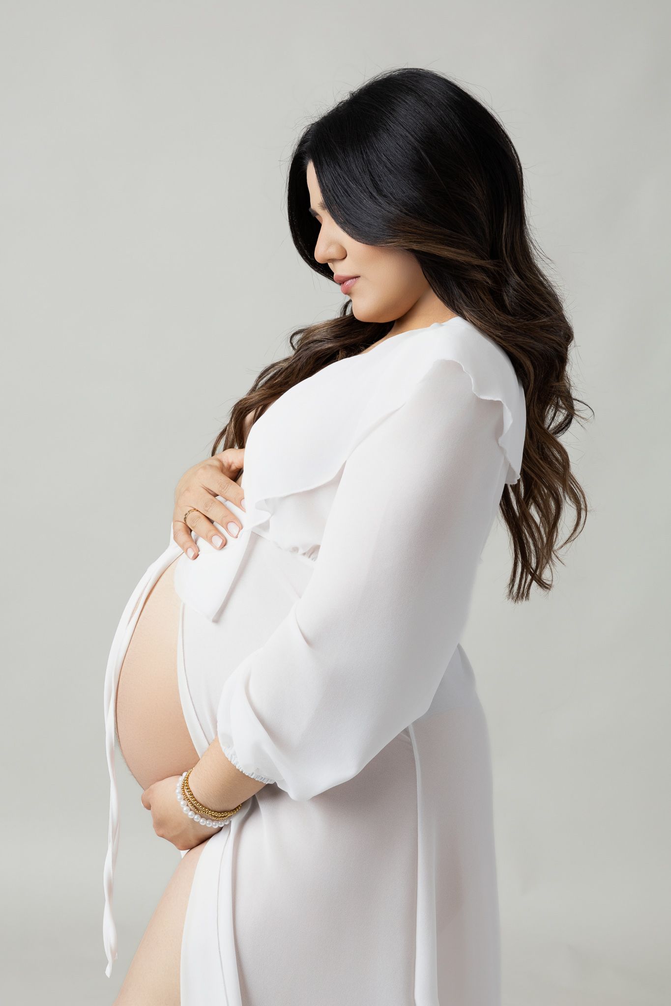 Elegant maternity portrait of woman in white robe against grey background.