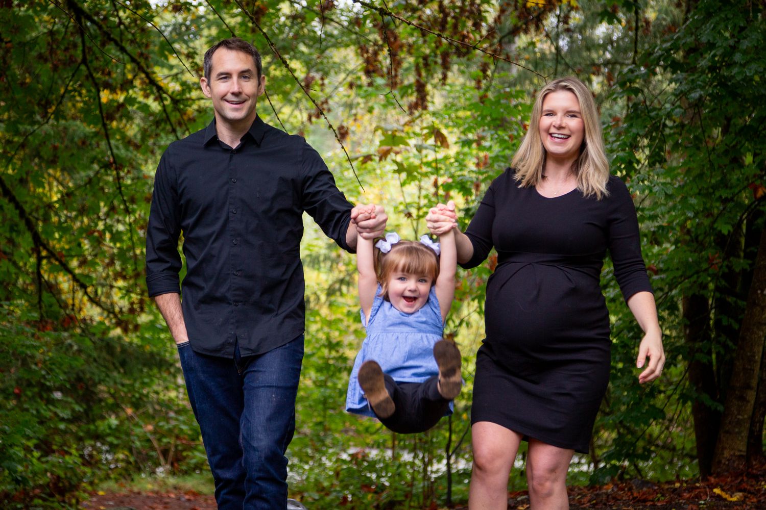 Family of three sharing candid laughter during outdoor photography session among green trees.