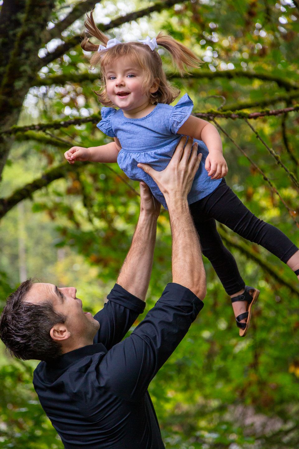 Joyful playtime moment captures child being lifted high in the air against lush green foliage backdrop.