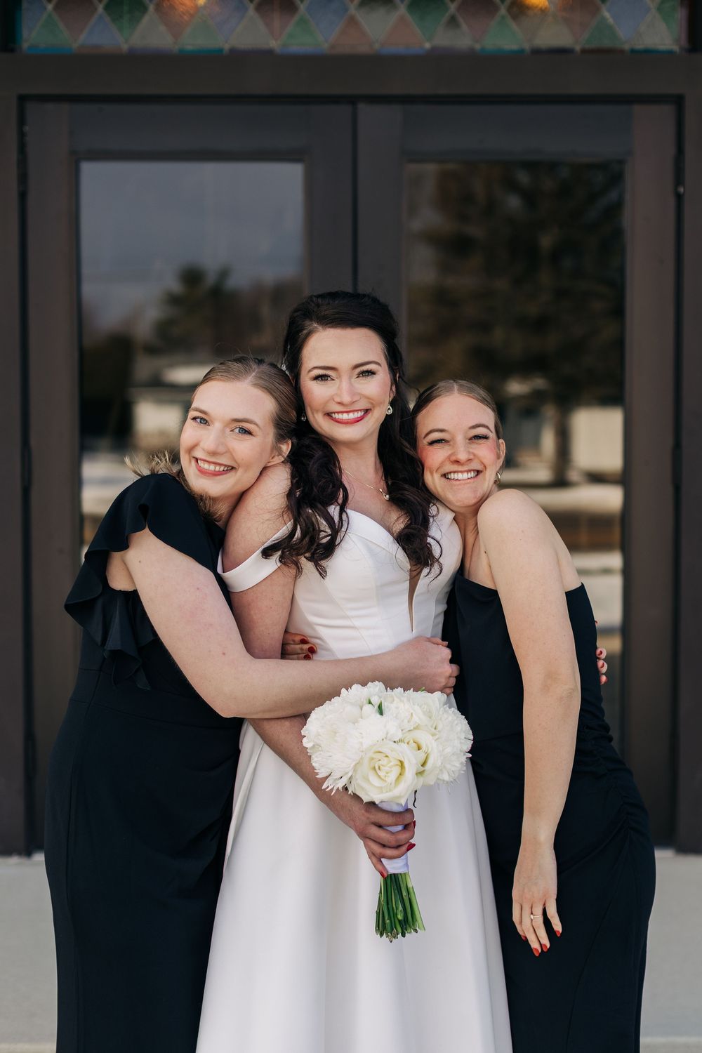 Three friends pose together at a wedding, with bridesmaids in black dresses hugging the bride in white holding a bouquet.